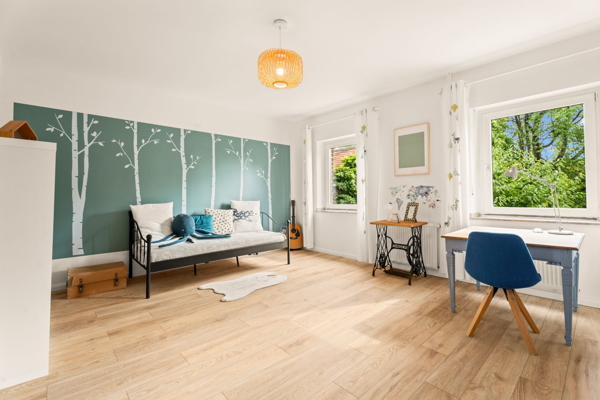 Helles Zimmer mit Holzboden, Bett, Schreibtisch und Wanddeko mit Baum-Motiv.