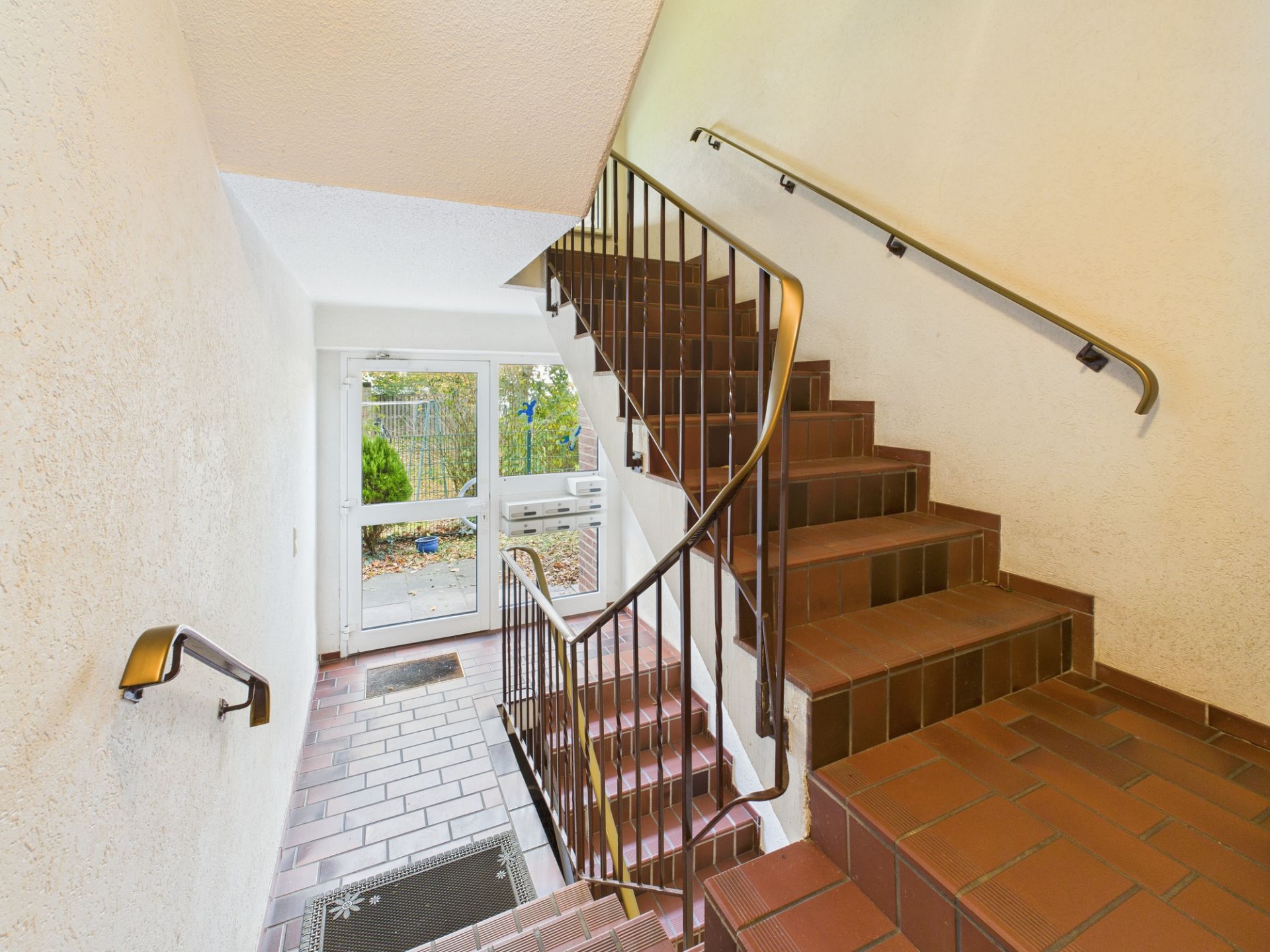Treppenhaus mit braunen Fliesen, Geländer und Glastür zum Garten.