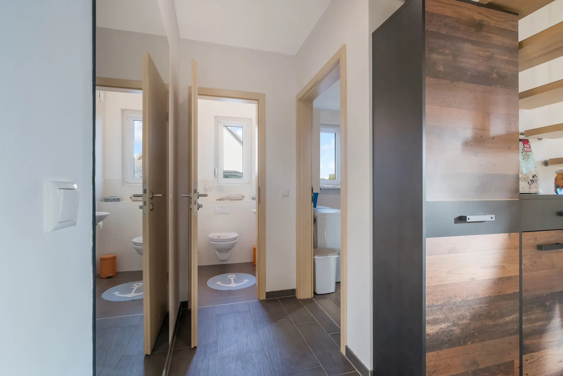 Flur mit Blick in zwei Bäder, eines mit Toilette und Fenster, modernes Holzdekor im Vordergrund.