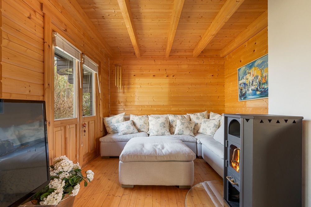 Wohnzimmer mit Holzwänden, Ecksofa, Kaminofen und großem Fenster.