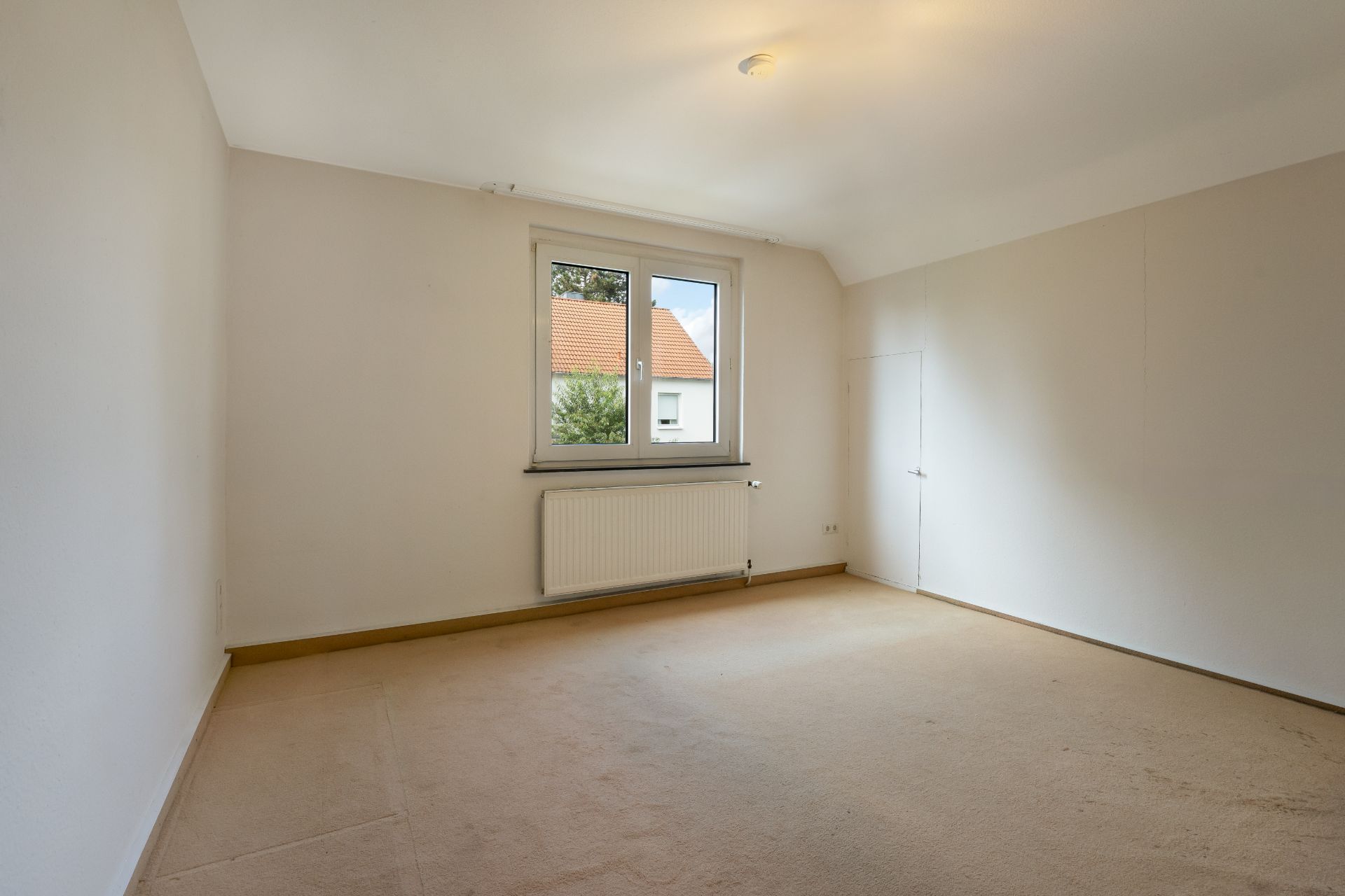 Leeres Zimmer mit Teppichboden, Fenster, Heizkörper und einer kleinen Tür an der rechten Wand.