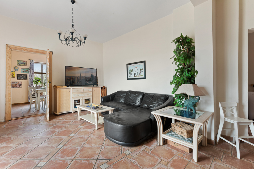 Wohnzimmer mit schwarzem Ledersofa, Fernseher, Couchtisch und Pflanze auf Fliesenboden.