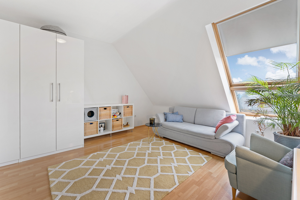 Helles Wohnzimmer mit Sofa, Sessel, Teppich, Schrank, Regal und großem Fenster mit Pflanze.