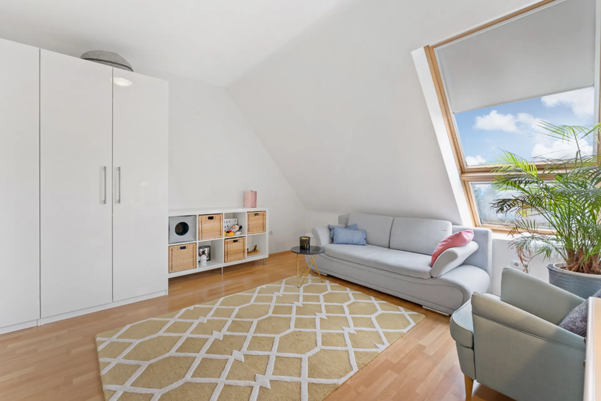 Helles Wohnzimmer mit Sofa, Sessel, Teppich, Schrank, Regal und großem Fenster mit Pflanze.