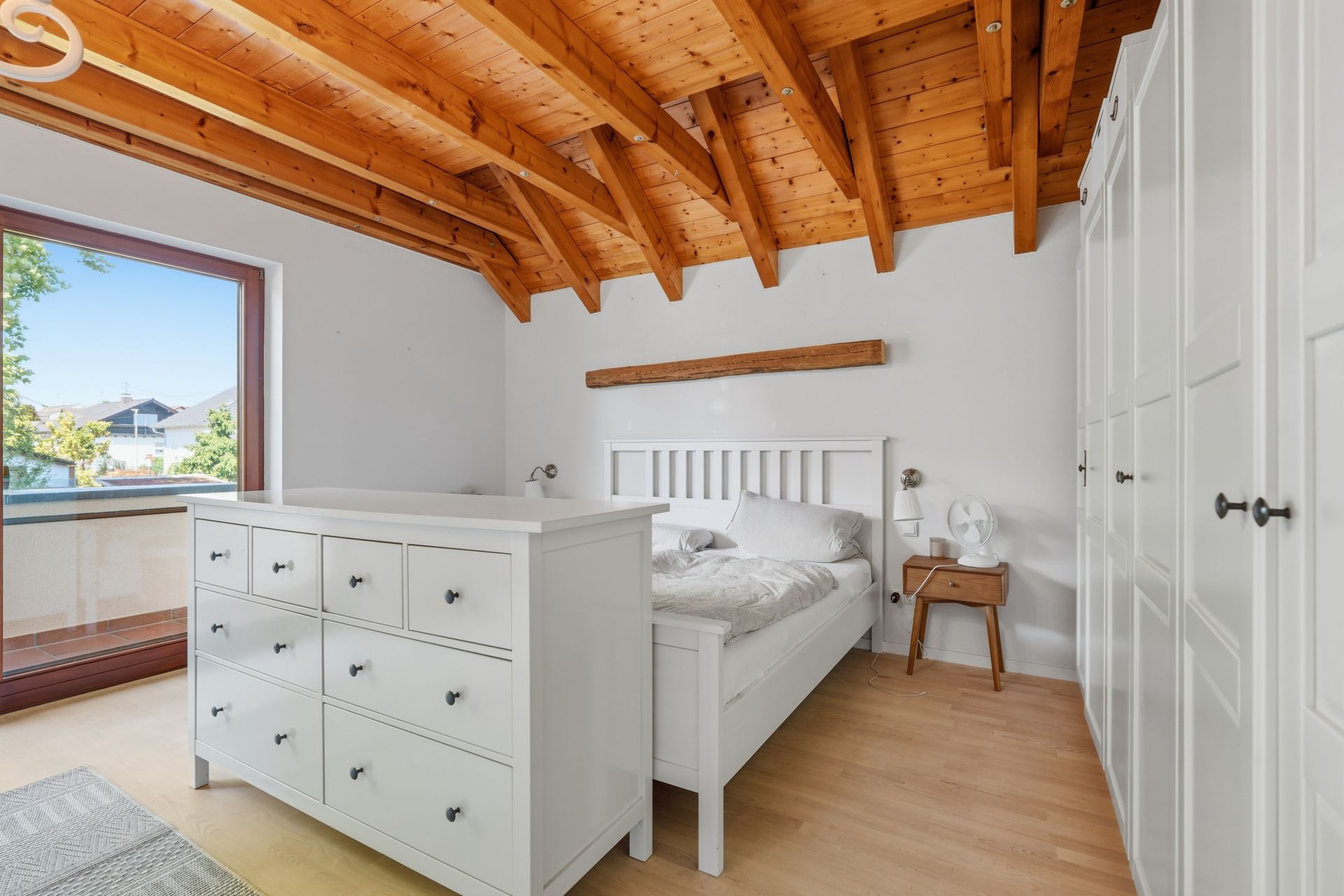 Helles Schlafzimmer mit Holzbalkendecke, weißem Bett, Kommode und großem Fenster.