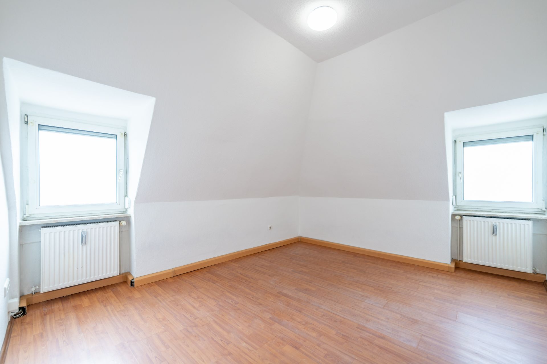 Helles, leeres Zimmer mit zwei Fenstern, Holzfußboden und weißen Wänden.