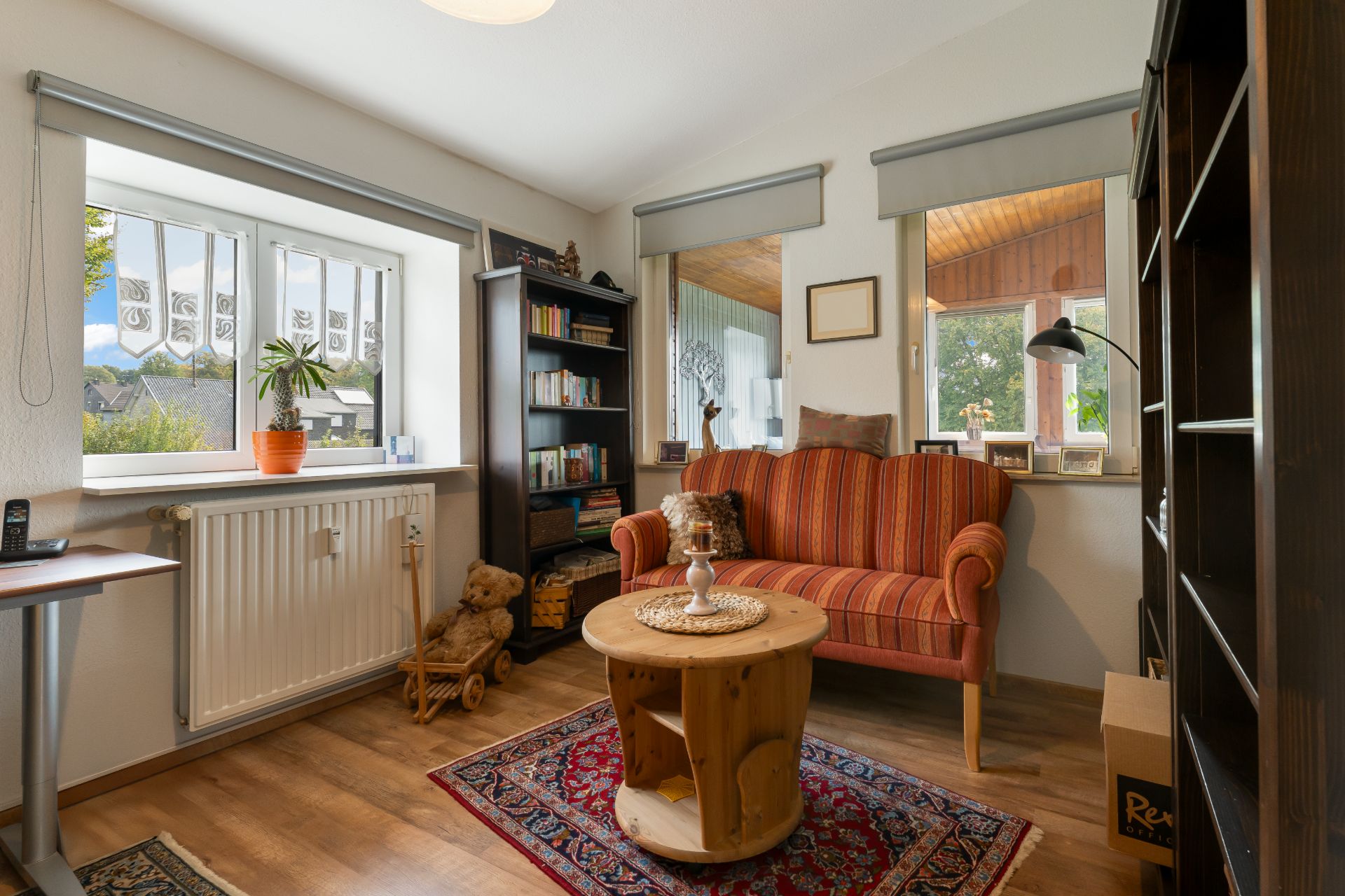 Helles Wohnzimmer mit rotem Sofa, Bücherregalen, kleinem Tisch und Fenstern.
