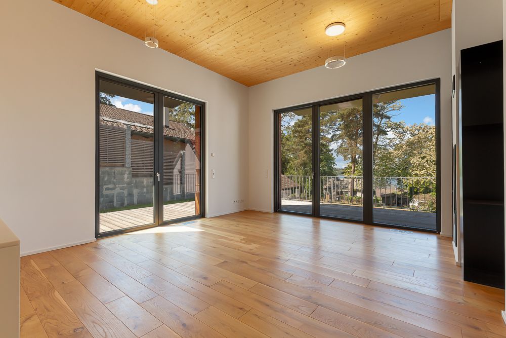 Helles Zimmer mit Holzfußboden, großen Fenstern und Blick auf Bäume und Terrasse.