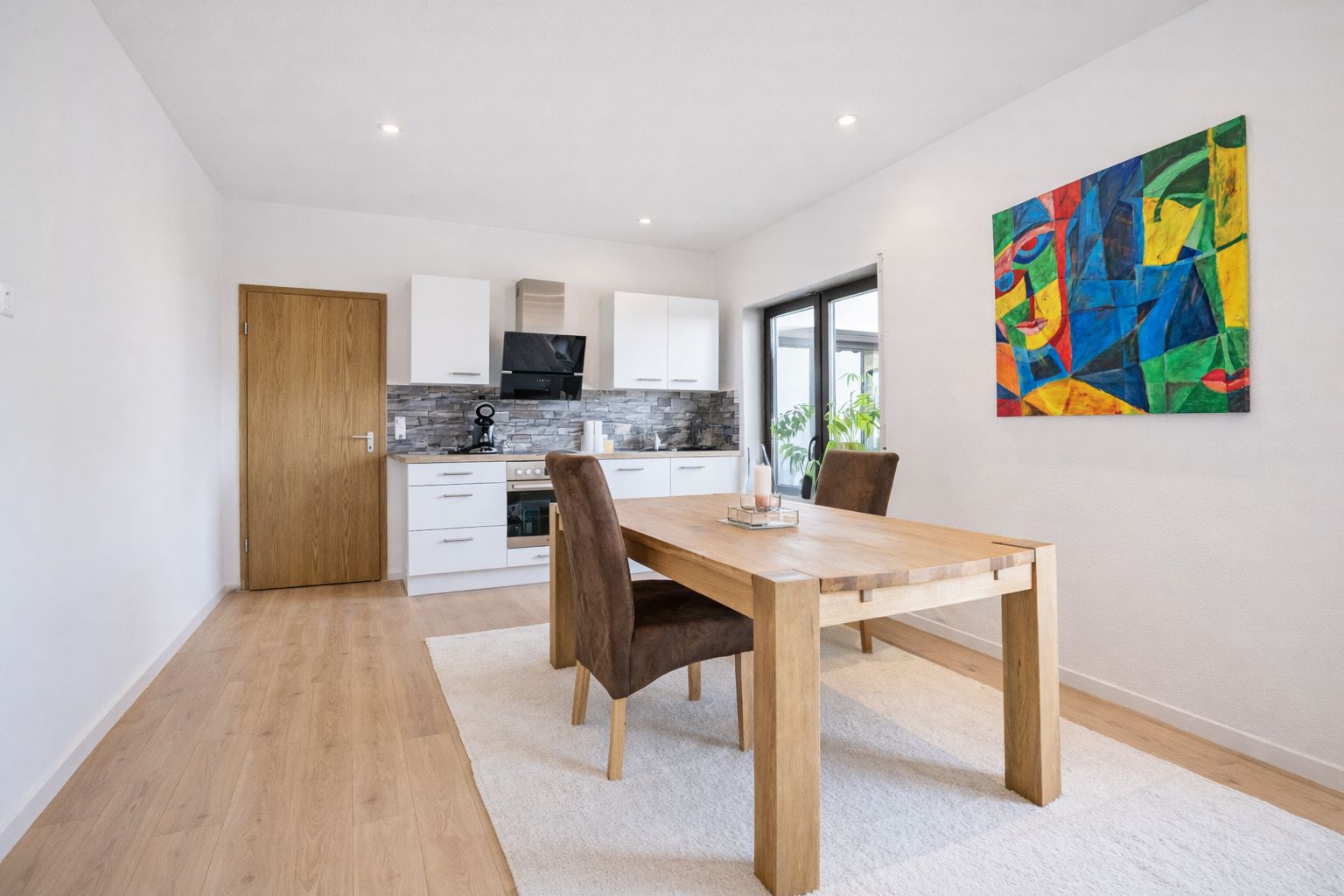 Moderne Küche mit Holztisch, zwei Stühlen, hellem Teppich und buntem Bild an der Wand.