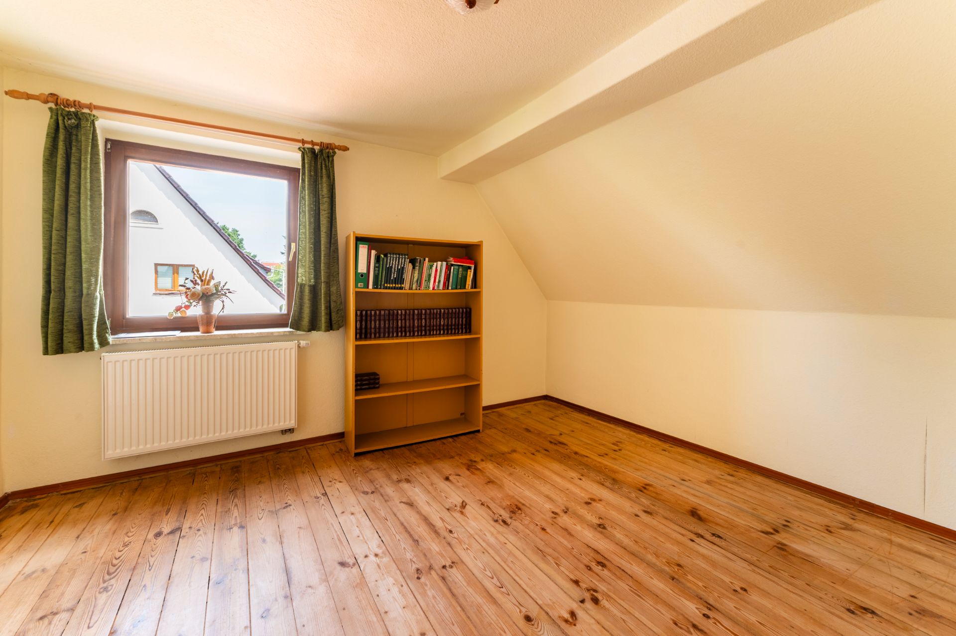 Helles Zimmer mit Holzboden, Fenster mit grünen Vorhängen und einem Bücherregal an der Wand.
