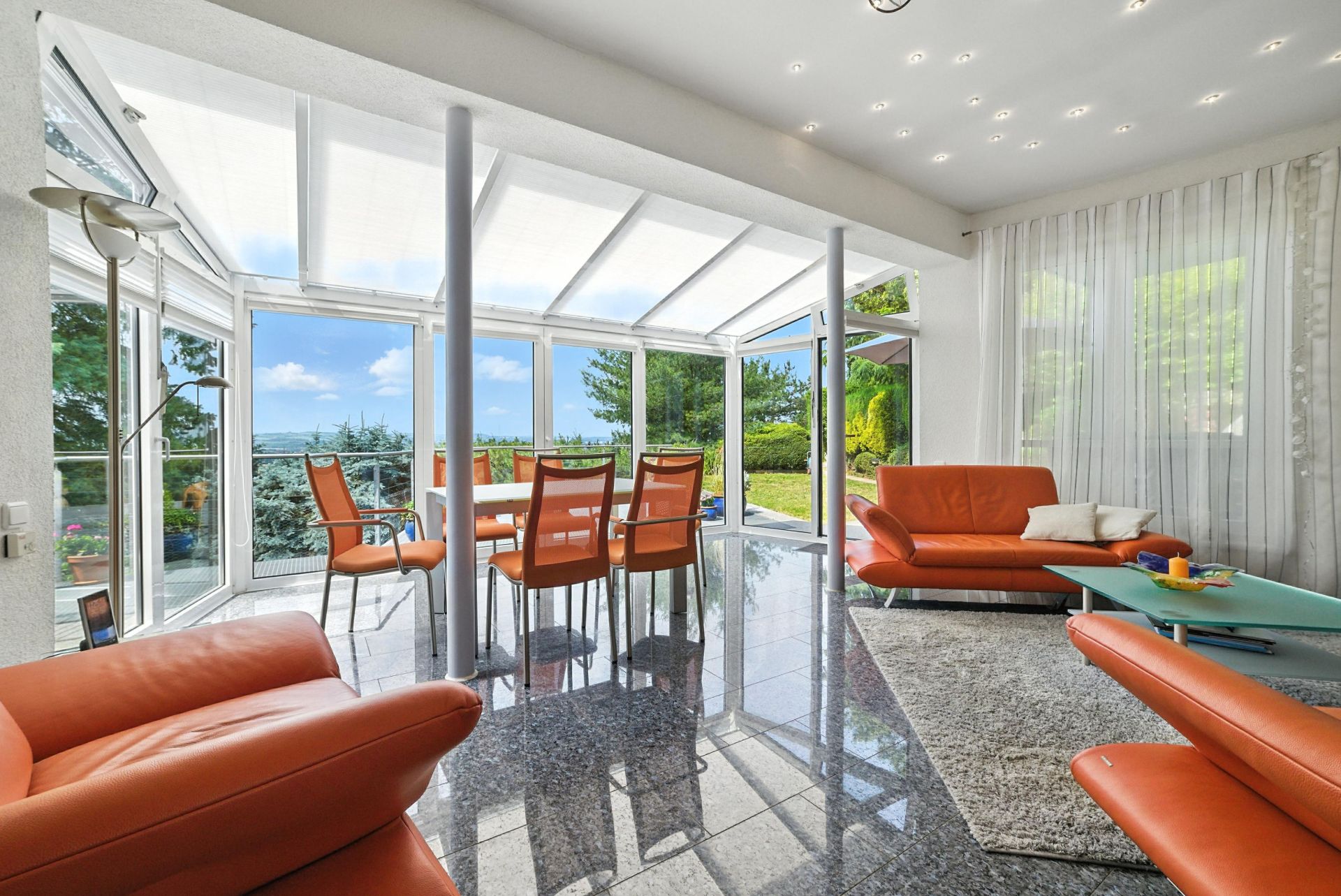 Wohnzimmer mit orangefarbenen Sofas, Esstisch, großen Fenstern und Blick in den Garten.