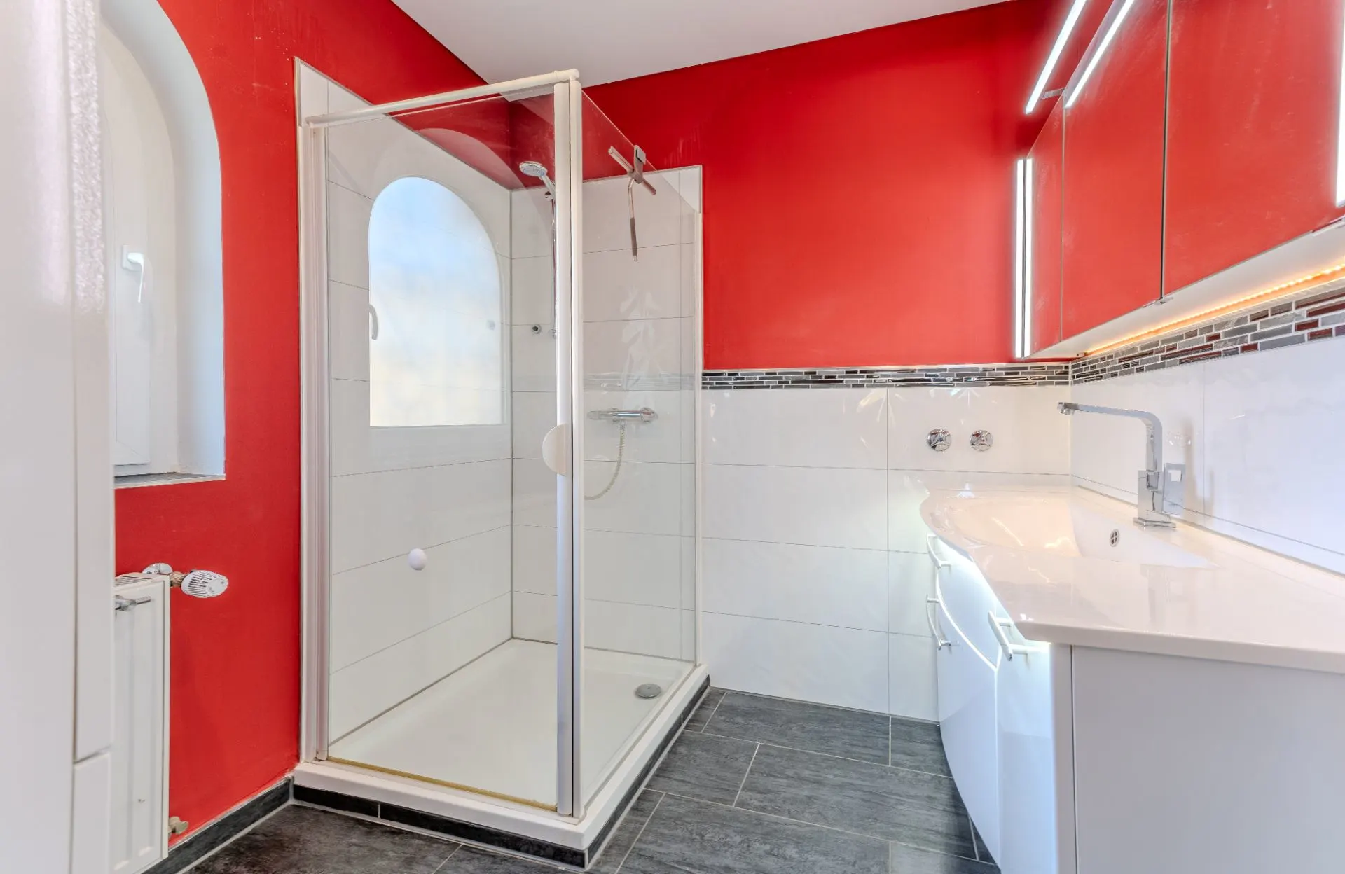 Modernes Badezimmer mit Dusche, roter Wand, großem Spiegel und weißem Waschbecken.