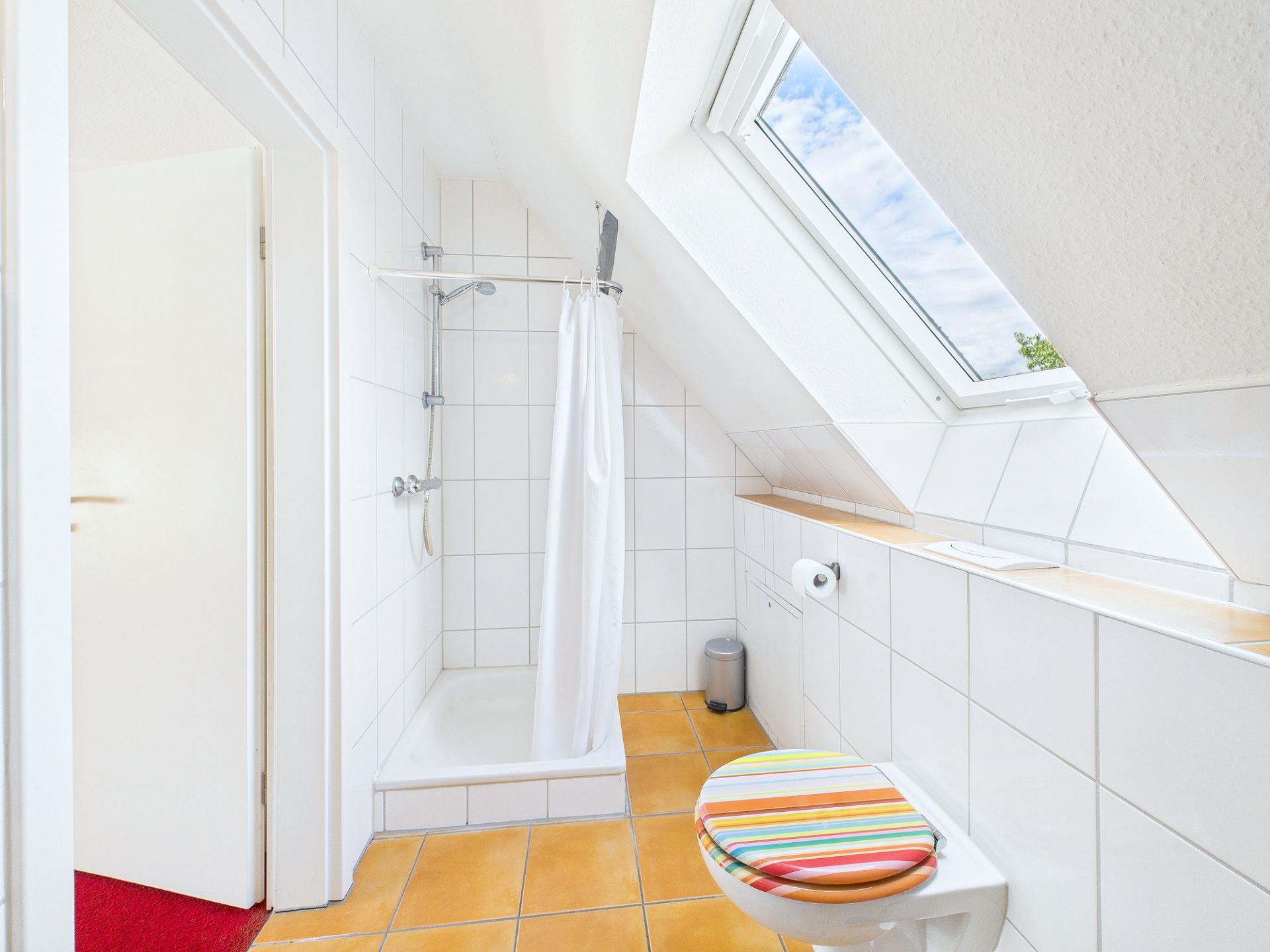 Helles Bad mit Dusche, WC und Dachfenster, gelbe Fliesen und bunter WC-Deckel.