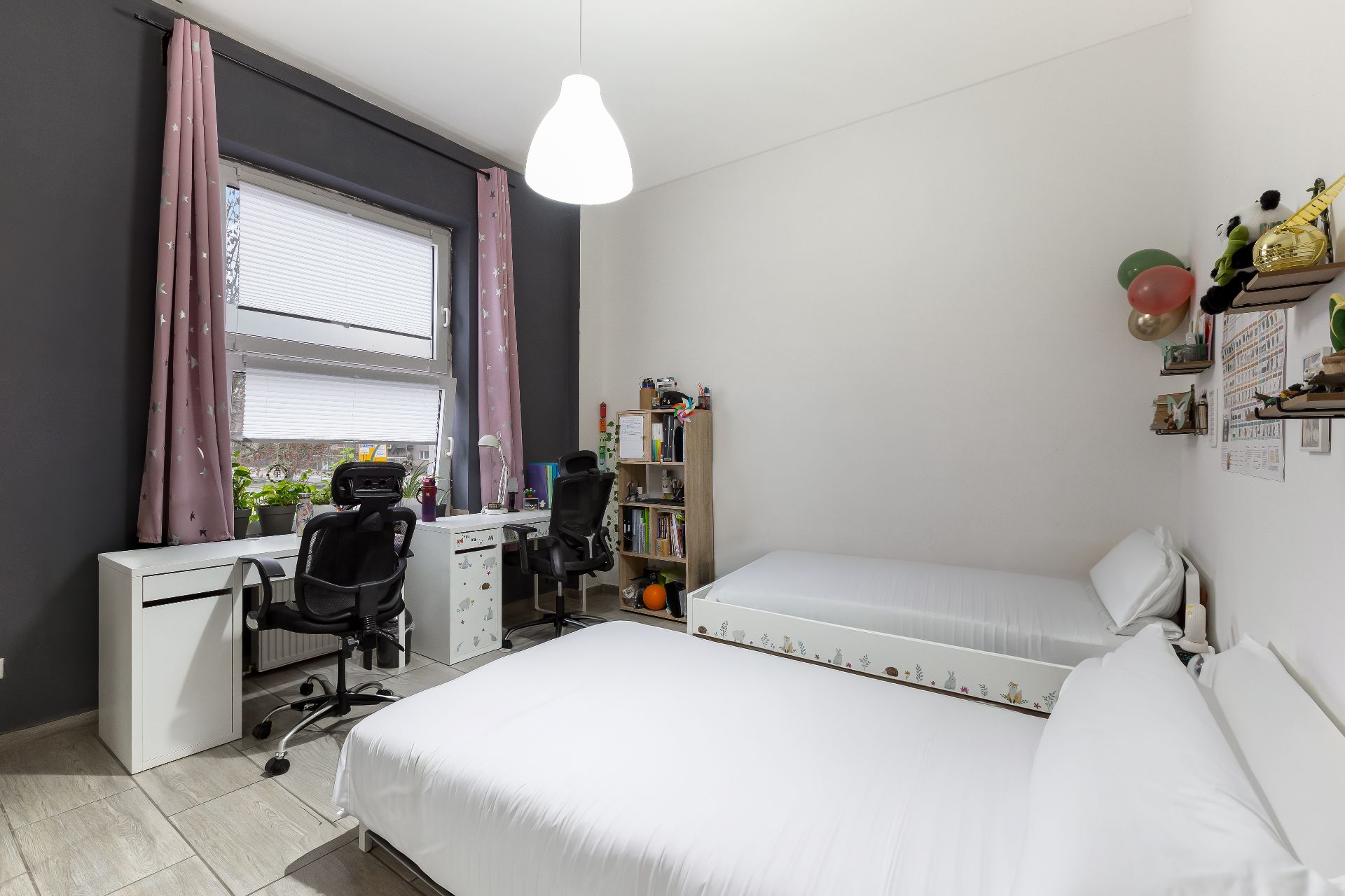 Helles Schlafzimmer mit zwei Betten, zwei Schreibtischen, Bücherregal und großem Fenster mit rosa Vorhängen.