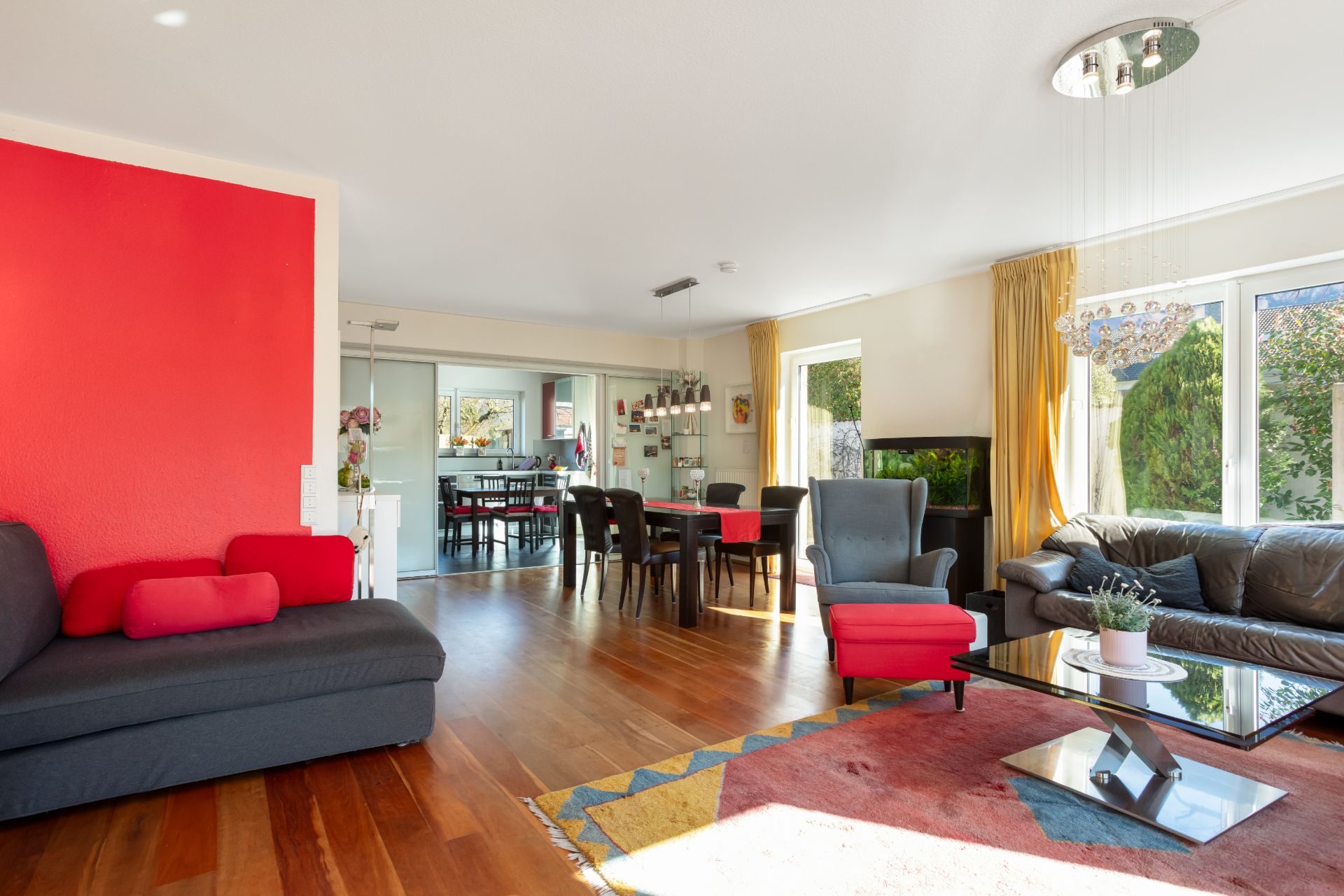 Helles Wohnzimmer mit roten Akzenten, Sofa, Sesseln, Esstisch und großen Fenstern zum Garten.