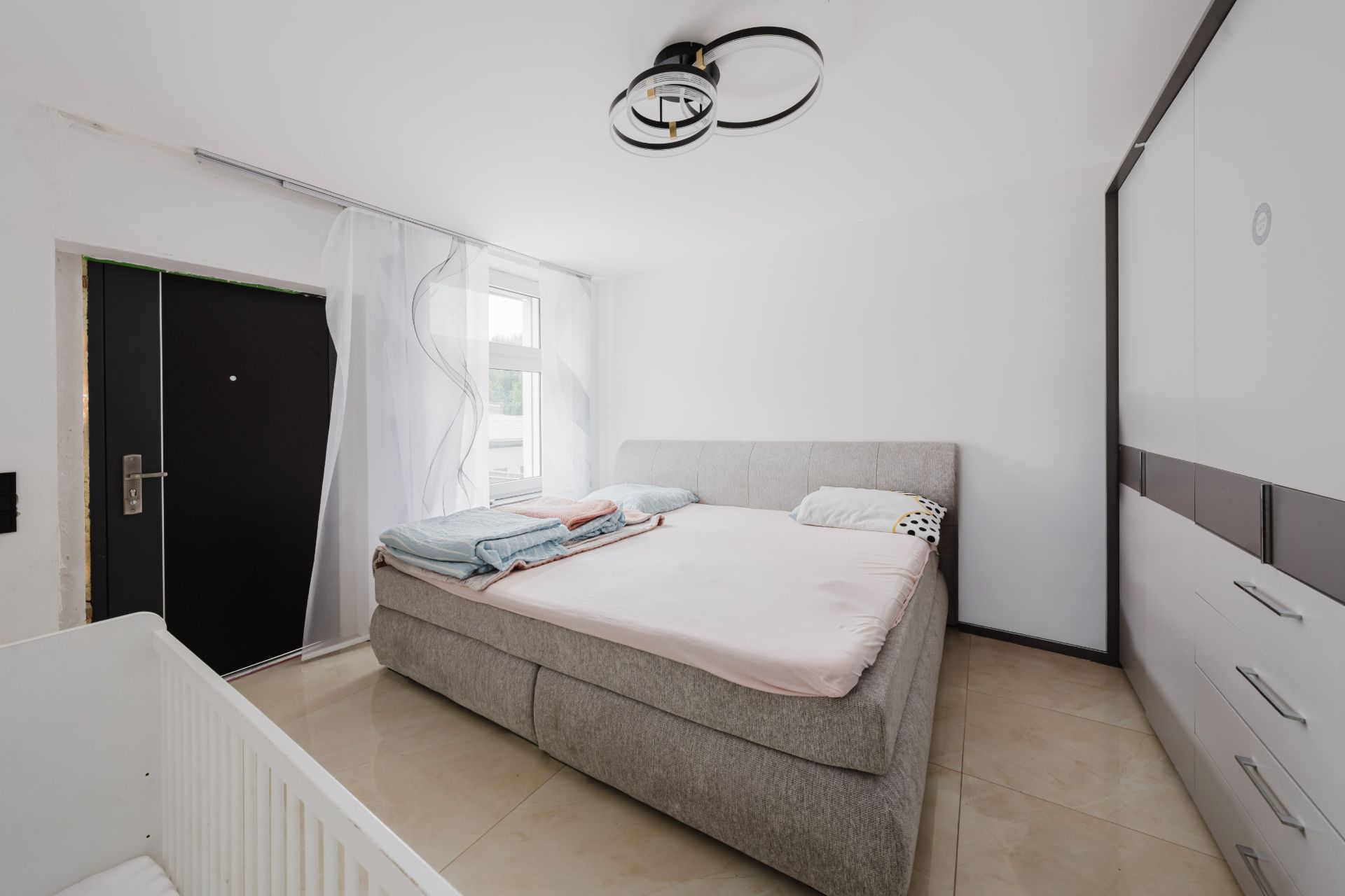 Schlafzimmer mit Doppelbett, Kleiderschrank, Babybett und Fenster mit weißen Vorhängen.