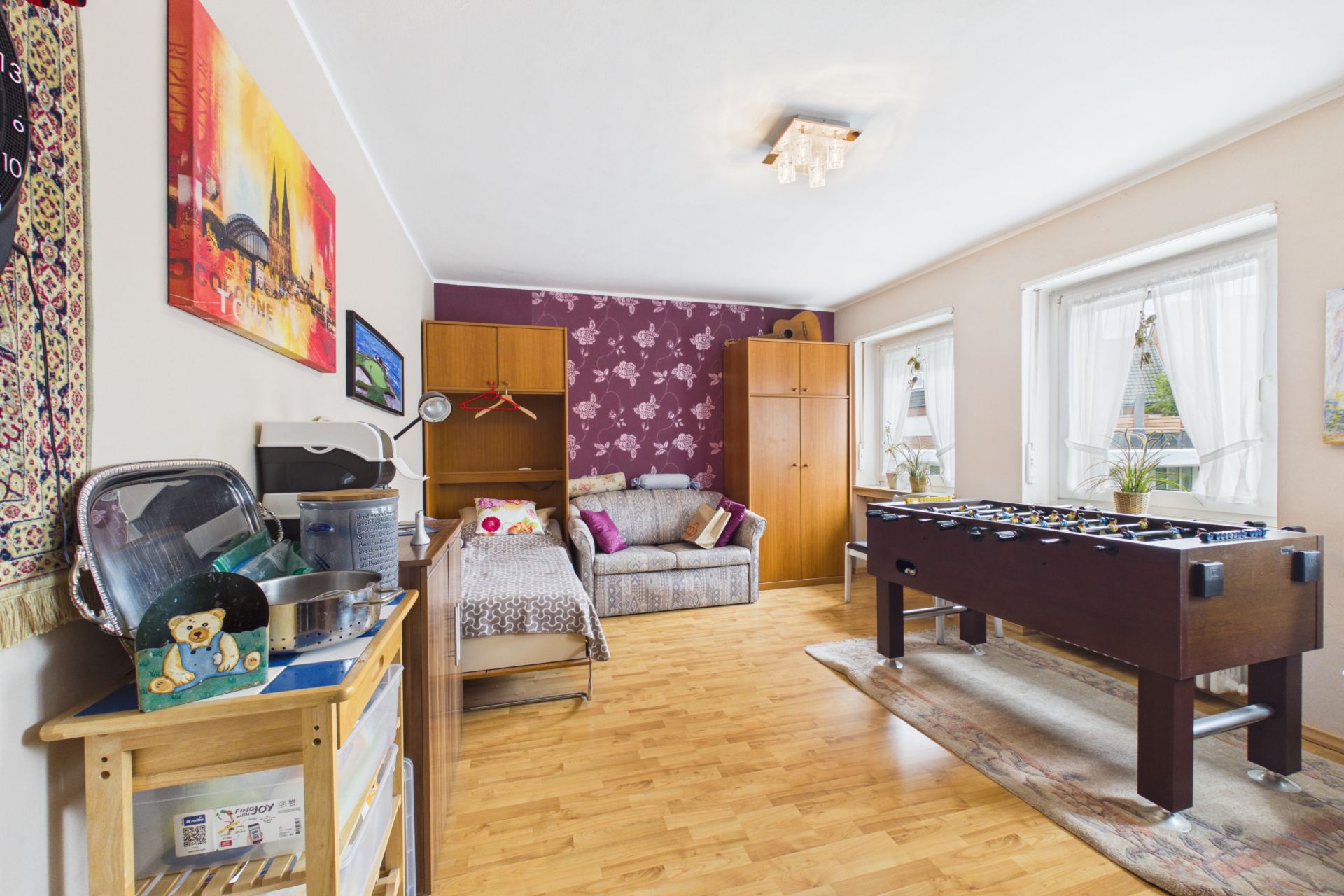Wohnzimmer mit Sofa, Schrank, Tischkicker und hellen Fenstern auf Holzfußboden.
