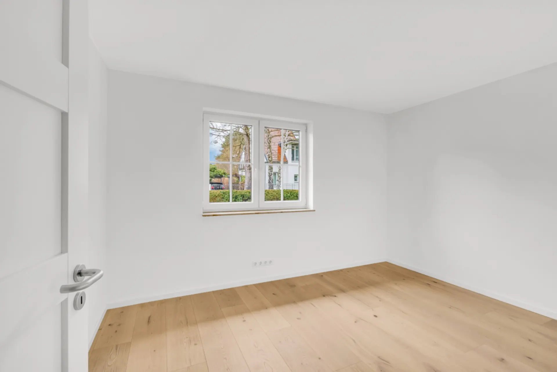 Leerer, heller Raum mit Holzboden und weiß gestrichenen Wänden, Fenster mit Blick nach draußen.
