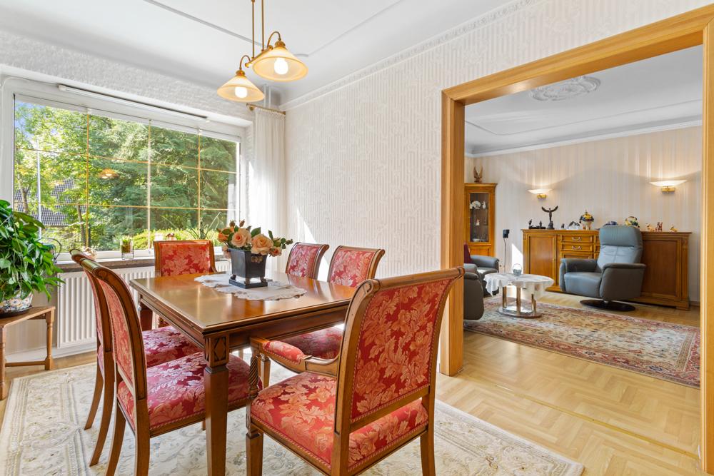 Esszimmer mit Holztisch, roten Stühlen und Blick ins Wohnzimmer mit Teppich und Sesseln.