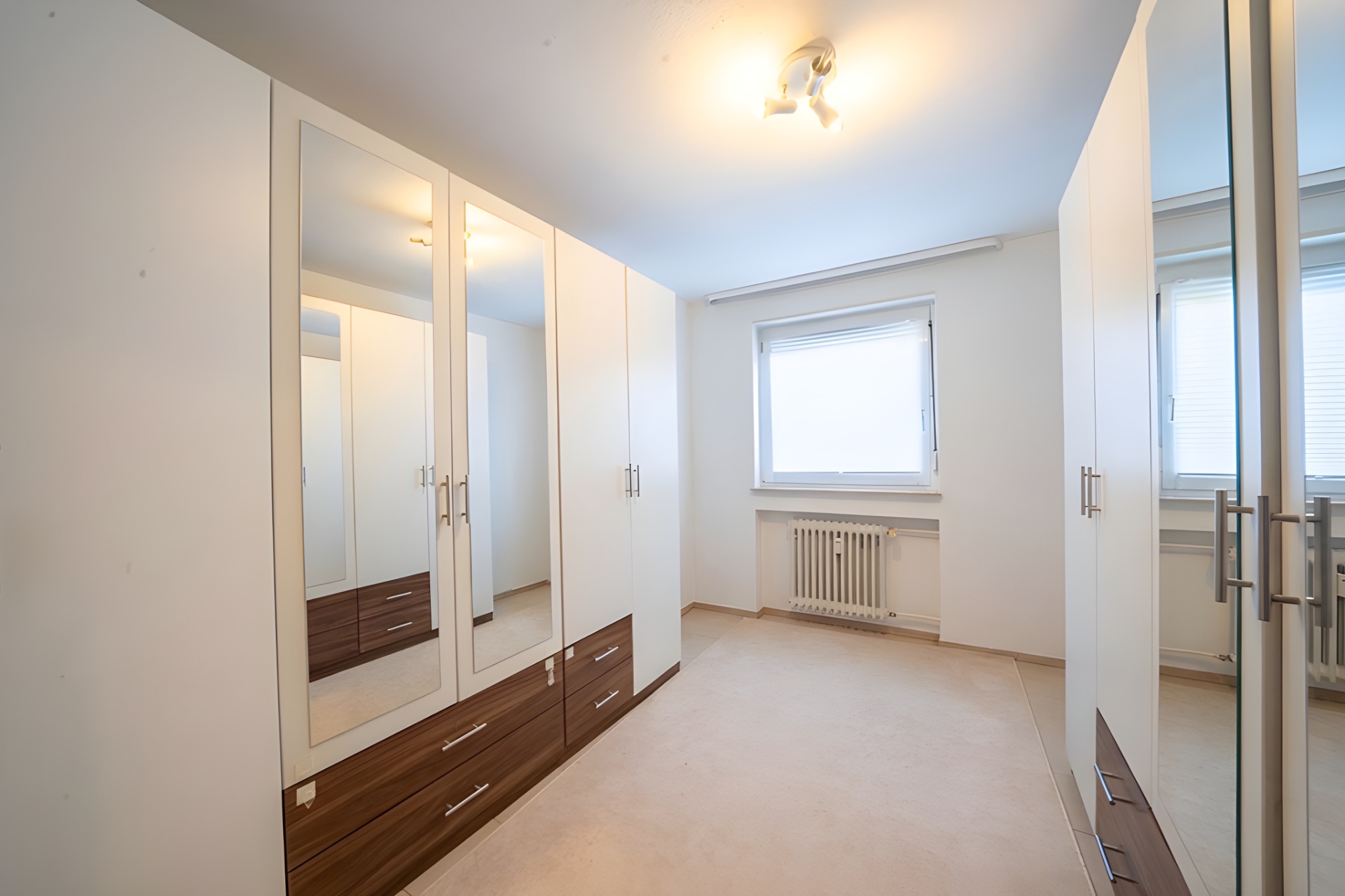 Heller Raum mit weißen Einbauschränken, Spiegeln und Fenster mit Rollladen.