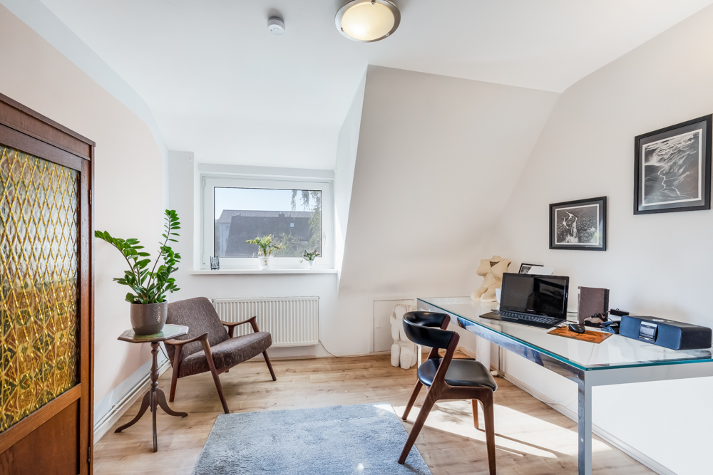 Helles Arbeitszimmer mit Glas-Schreibtisch, Stuhl, Sessel, Pflanze und Bildern an der Wand.