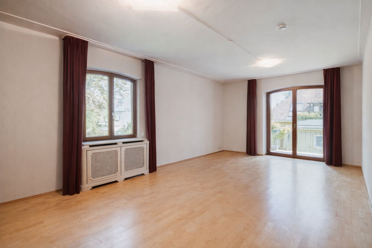 Heller, leerer Raum mit Holzfußboden, zwei Fenstern und braunen Vorhängen.