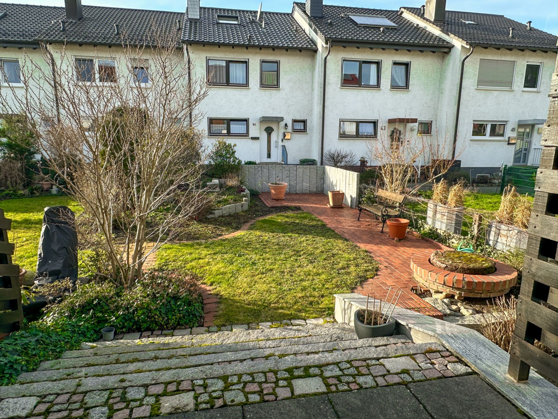 Rasengarten mit Terrasse, runden Beeten und Blick auf Reihenhäuser im Hintergrund.