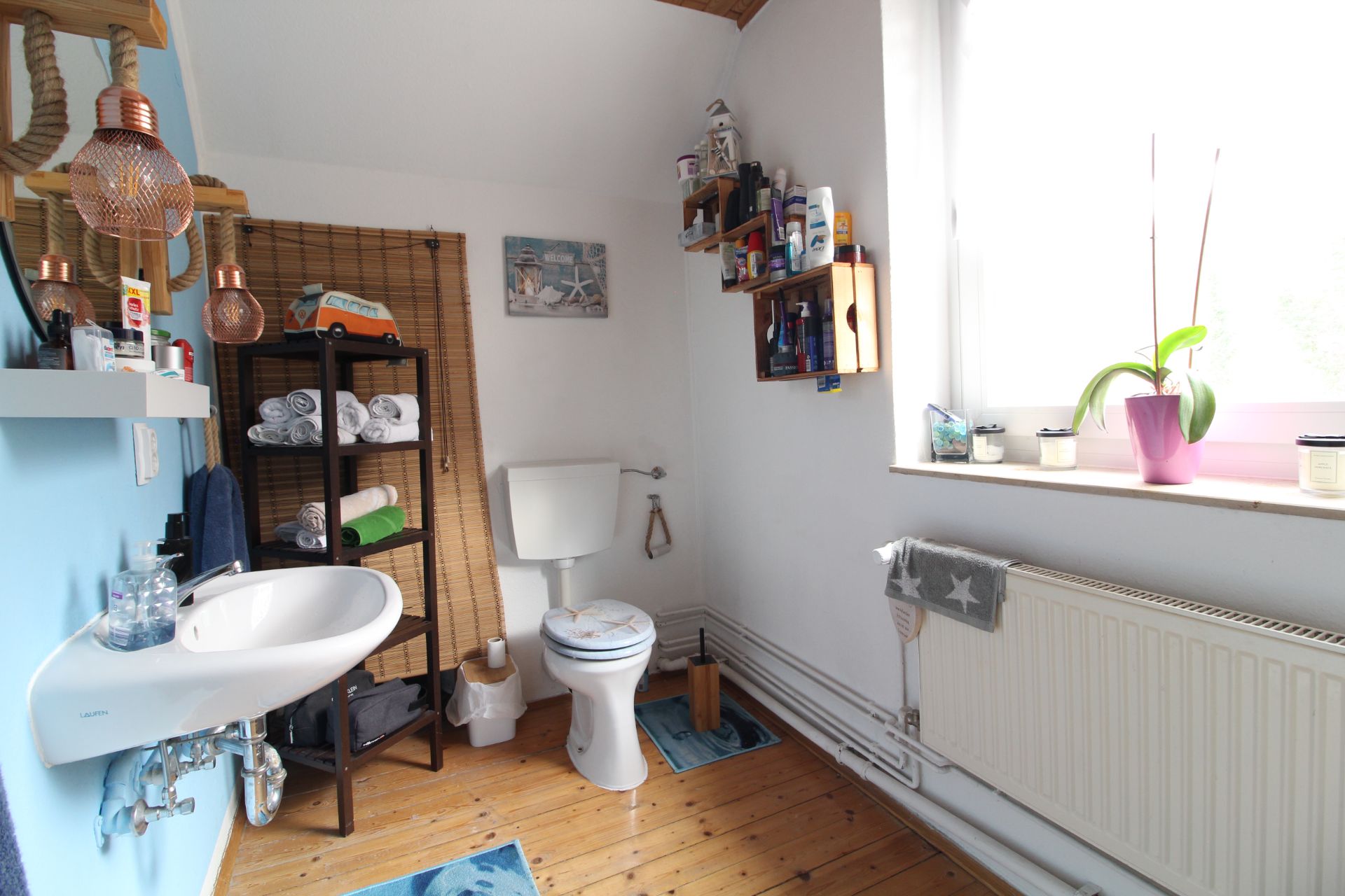 Helles Badezimmer mit Waschbecken, Toilette, Regal, Fenster und Pflanzen auf Holzfußboden.