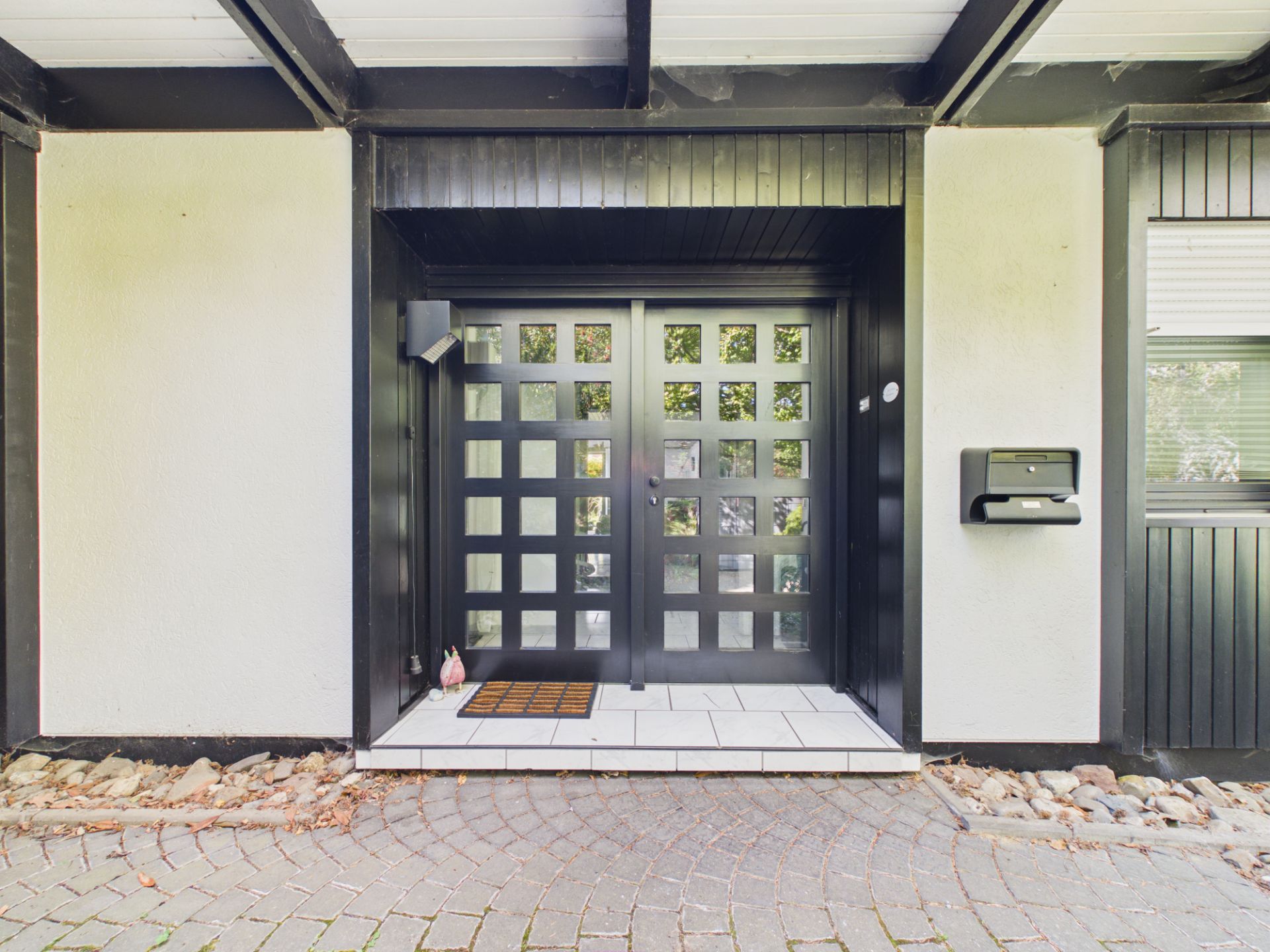 Eingangsbereich mit schwarzer Glastür, Fußmatte und Briefkasten an der Wand.