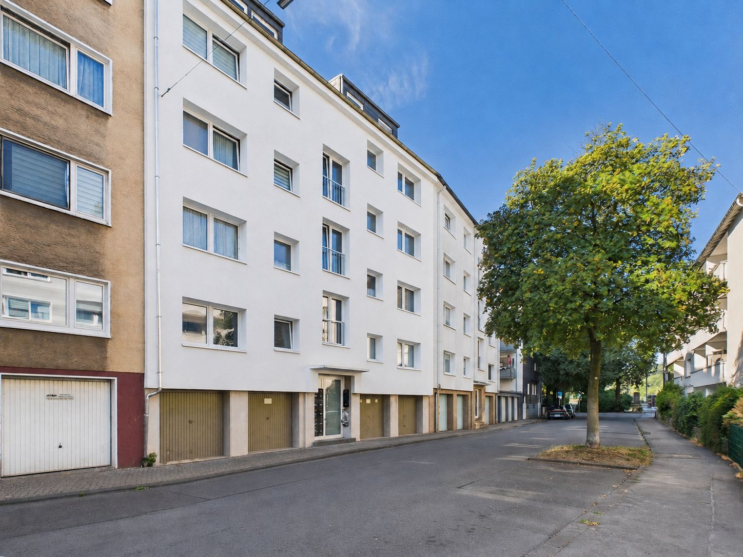 Mehrstöckiges Wohnhaus mit Garagen im Erdgeschoss, Baum am Straßenrand, blauer Himmel.