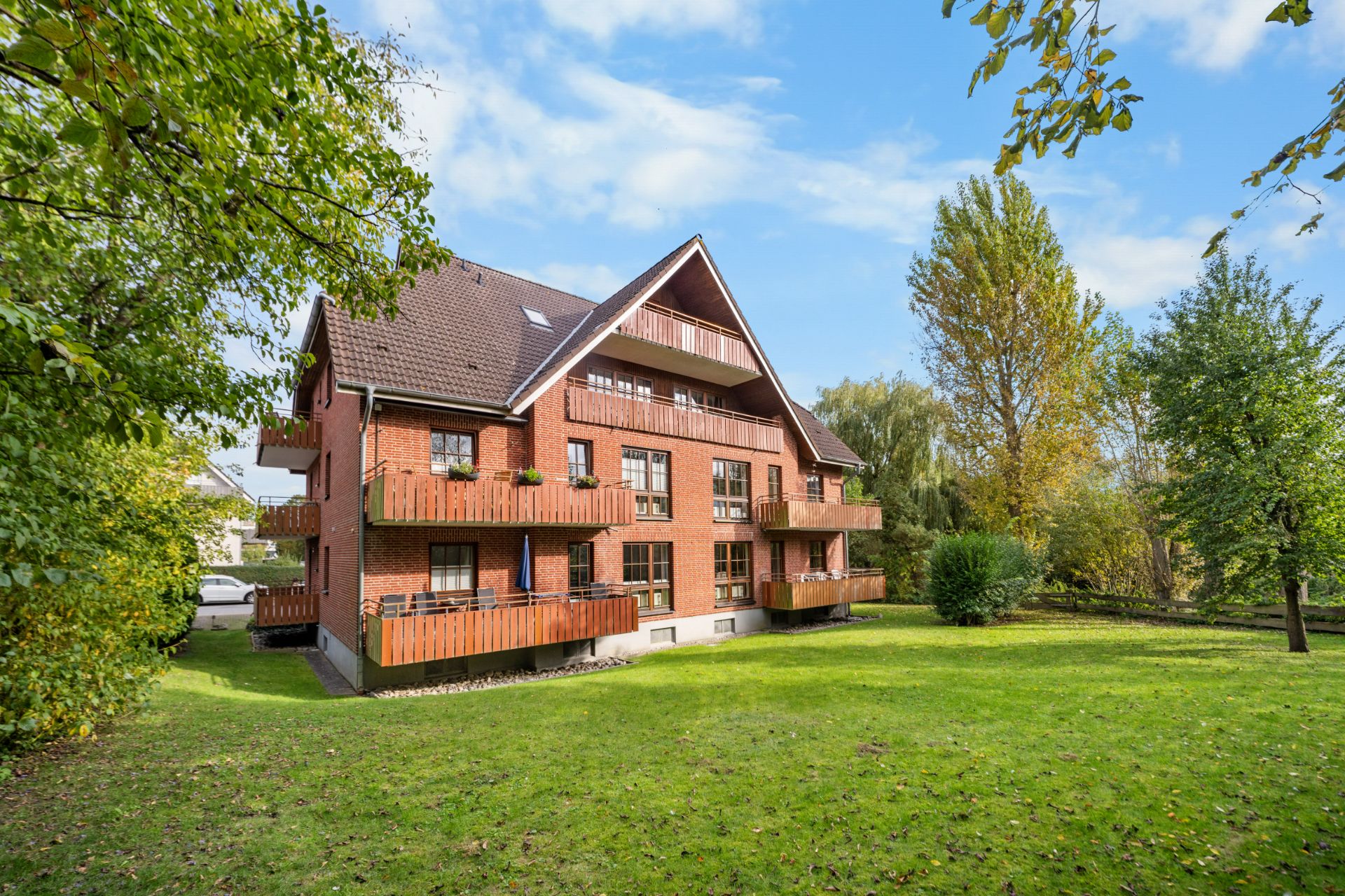 Rotes Mehrfamilienhaus mit Balkonen und großem Garten, umgeben von Bäumen bei sonnigem Wetter.