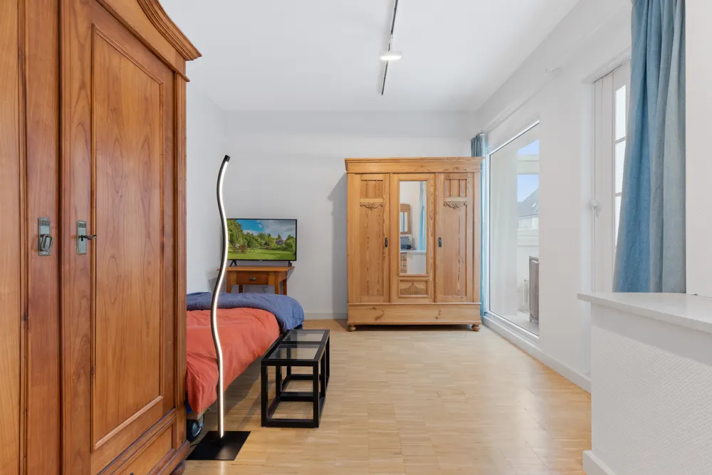 Helles Schlafzimmer mit zwei Holzschränken, Bett, Fernseher und großem Fenster mit blauen Vorhängen.