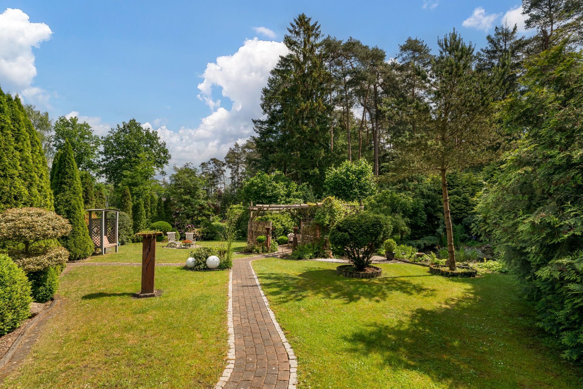 Gepflegter Garten mit Rasen, Bäumen, Sträuchern und einem gepflasterten Weg unter blauem Himmel.