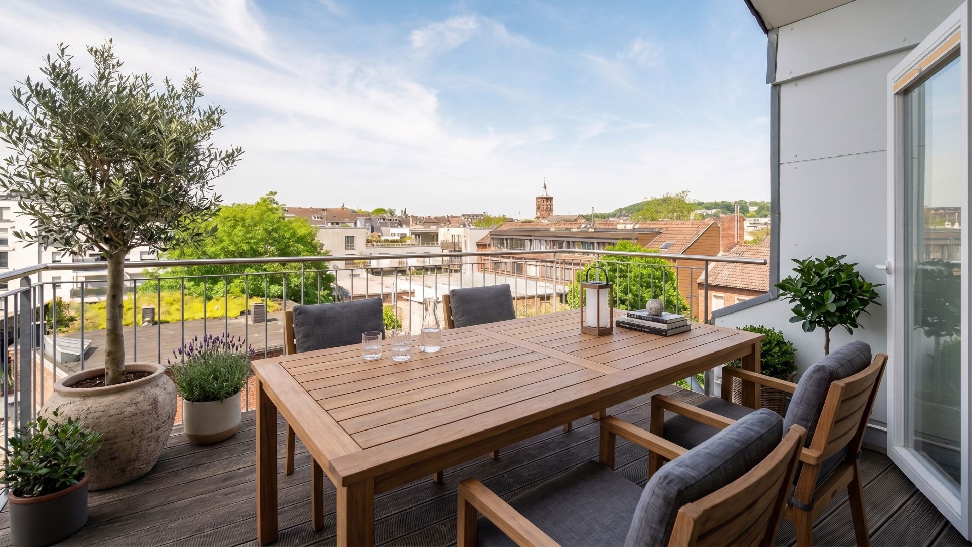 Balkon mit Holztisch, vier Stühlen, Pflanzen und Blick auf Stadt und Dächer.