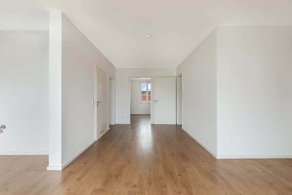 Helles, leeres Zimmer mit weiß gestrichenen Wänden und Holzfußboden, mehrere Türen sichtbar.