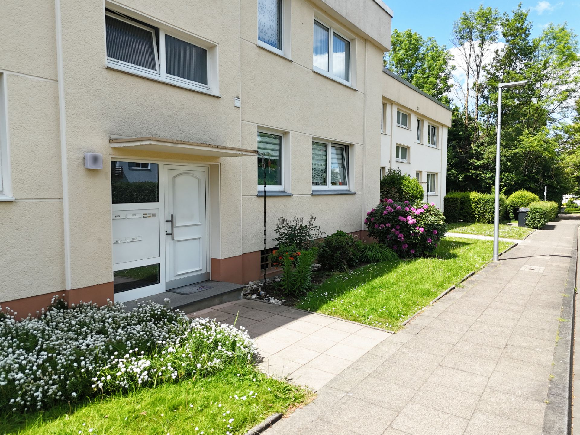Eingang eines Wohnhauses mit Blumenbeeten und Gehweg daneben.