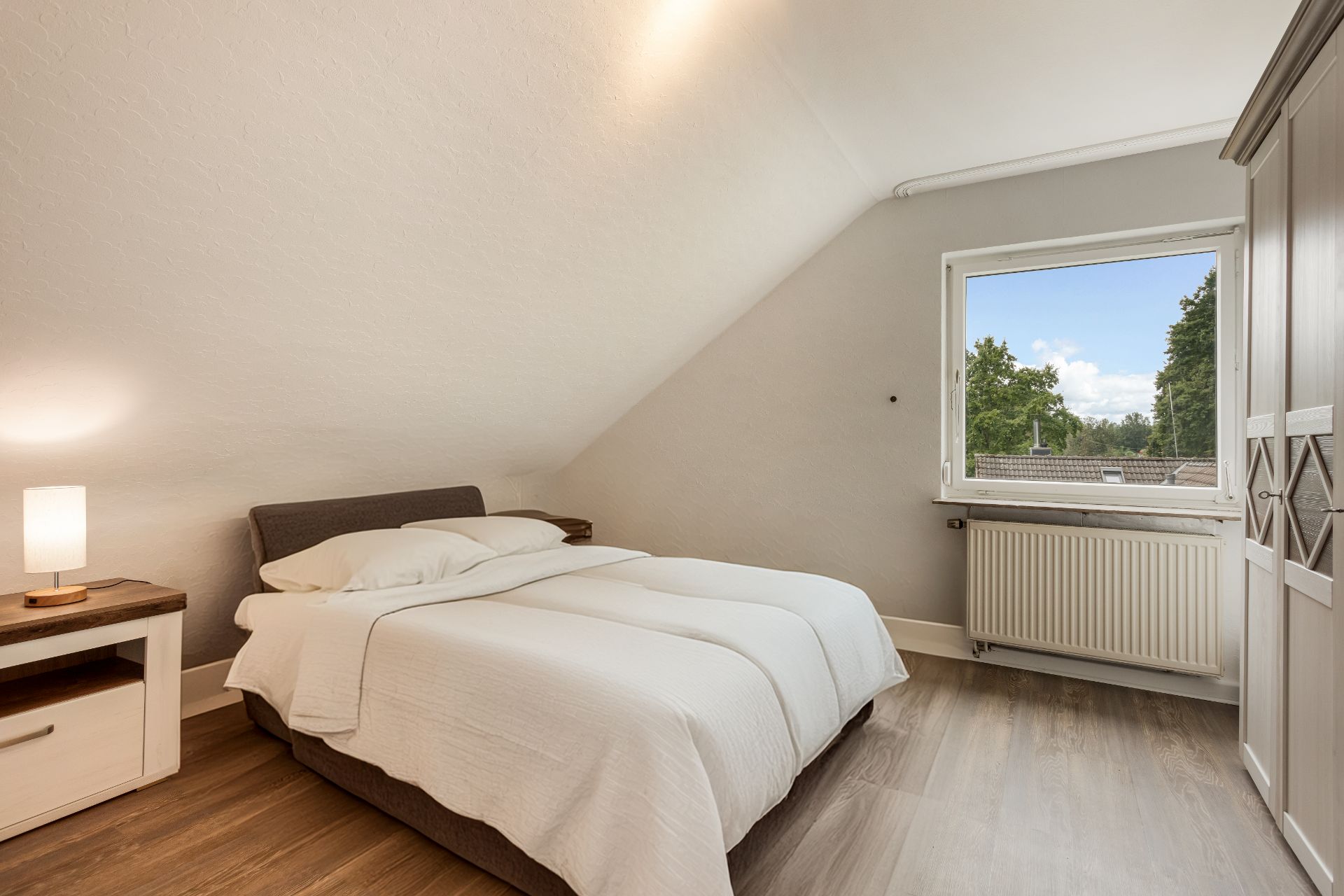 Helles Schlafzimmer mit Doppelbett, Nachttischlampe, Fenster und Kleiderschrank.