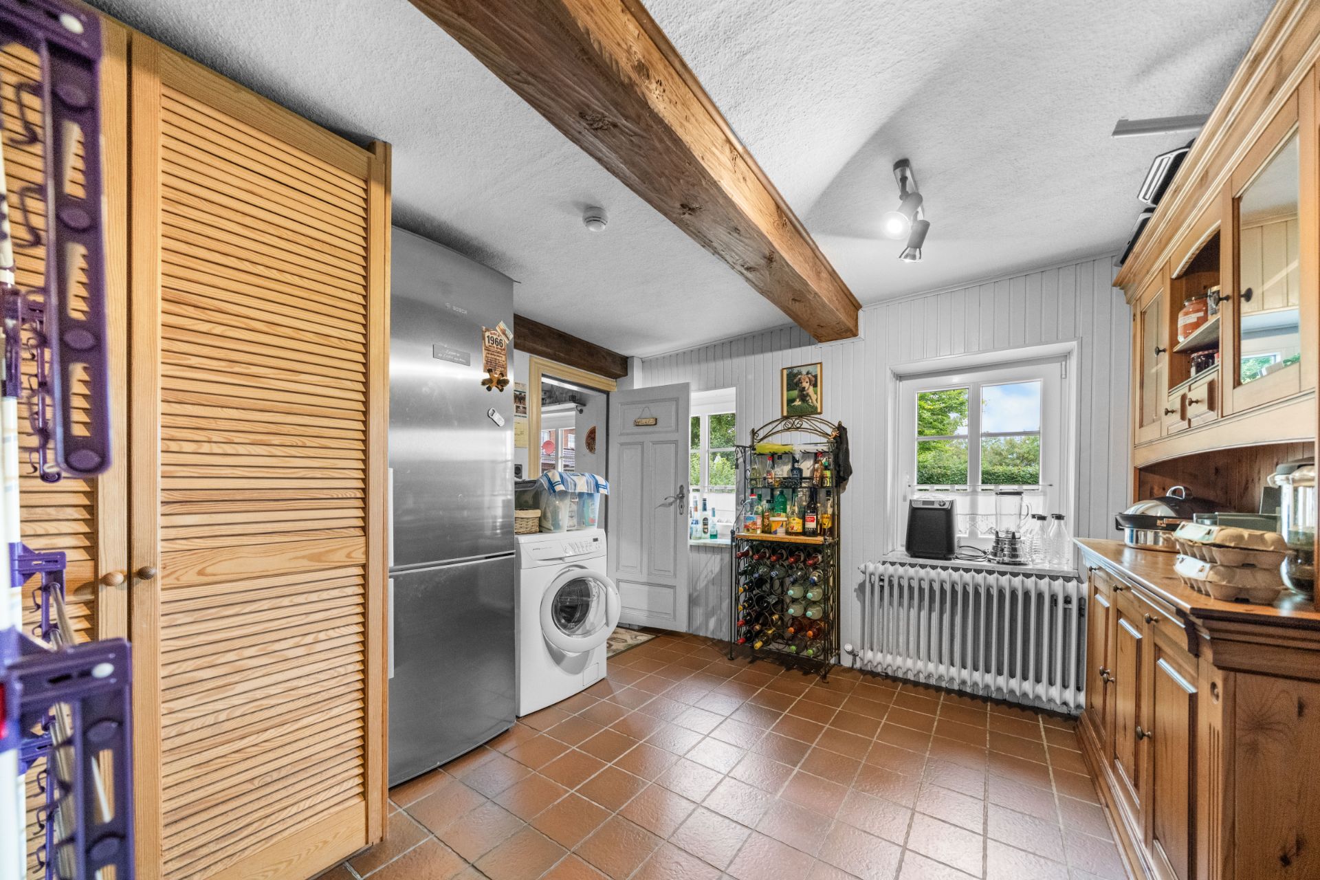 Helle Küche mit Fliesenboden, Kühlschrank, Waschmaschine, Holzschrank und Fenstern.