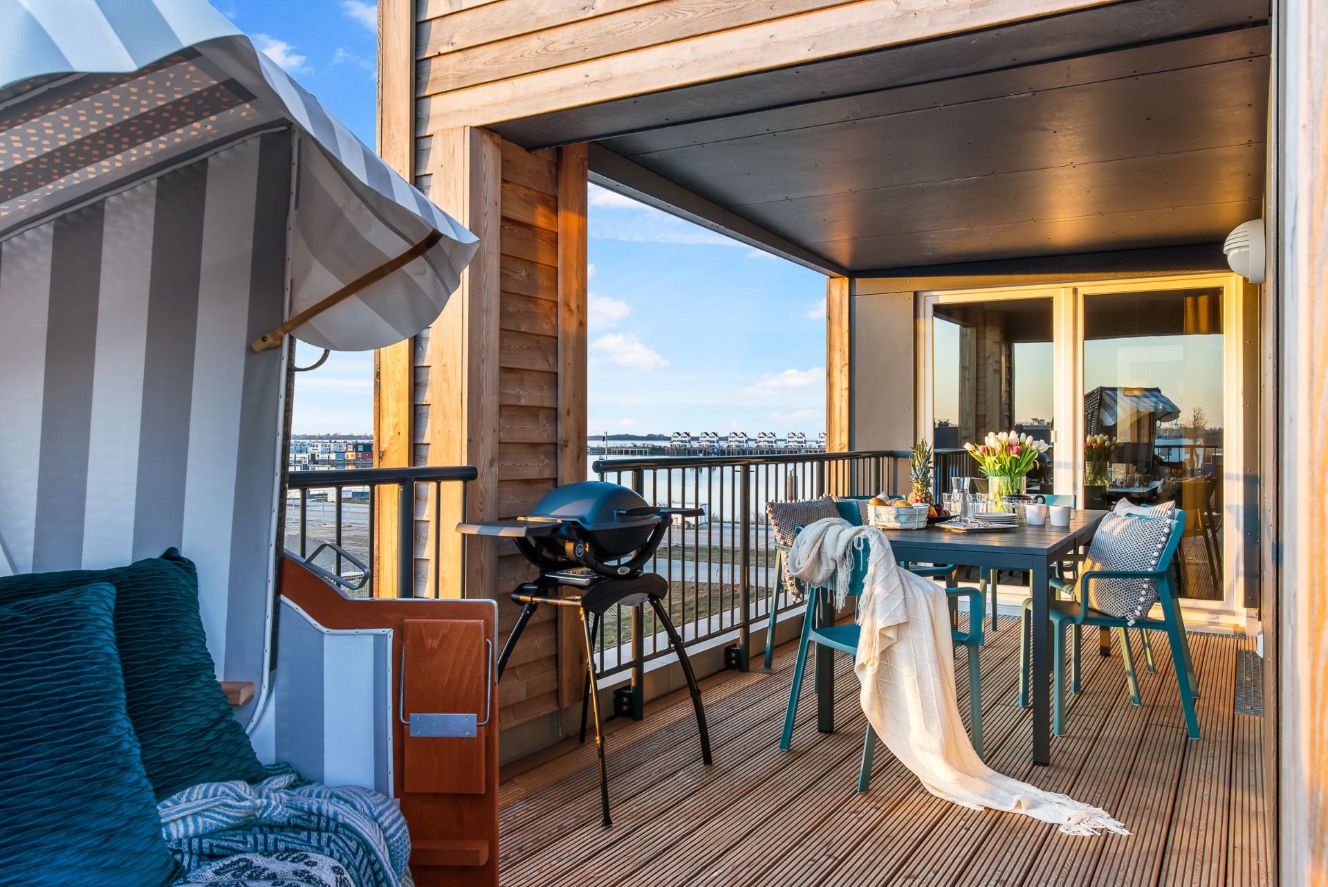 Überdachter Balkon mit Esstisch, Grill und Strandkorb, Blick auf das Wasser.
