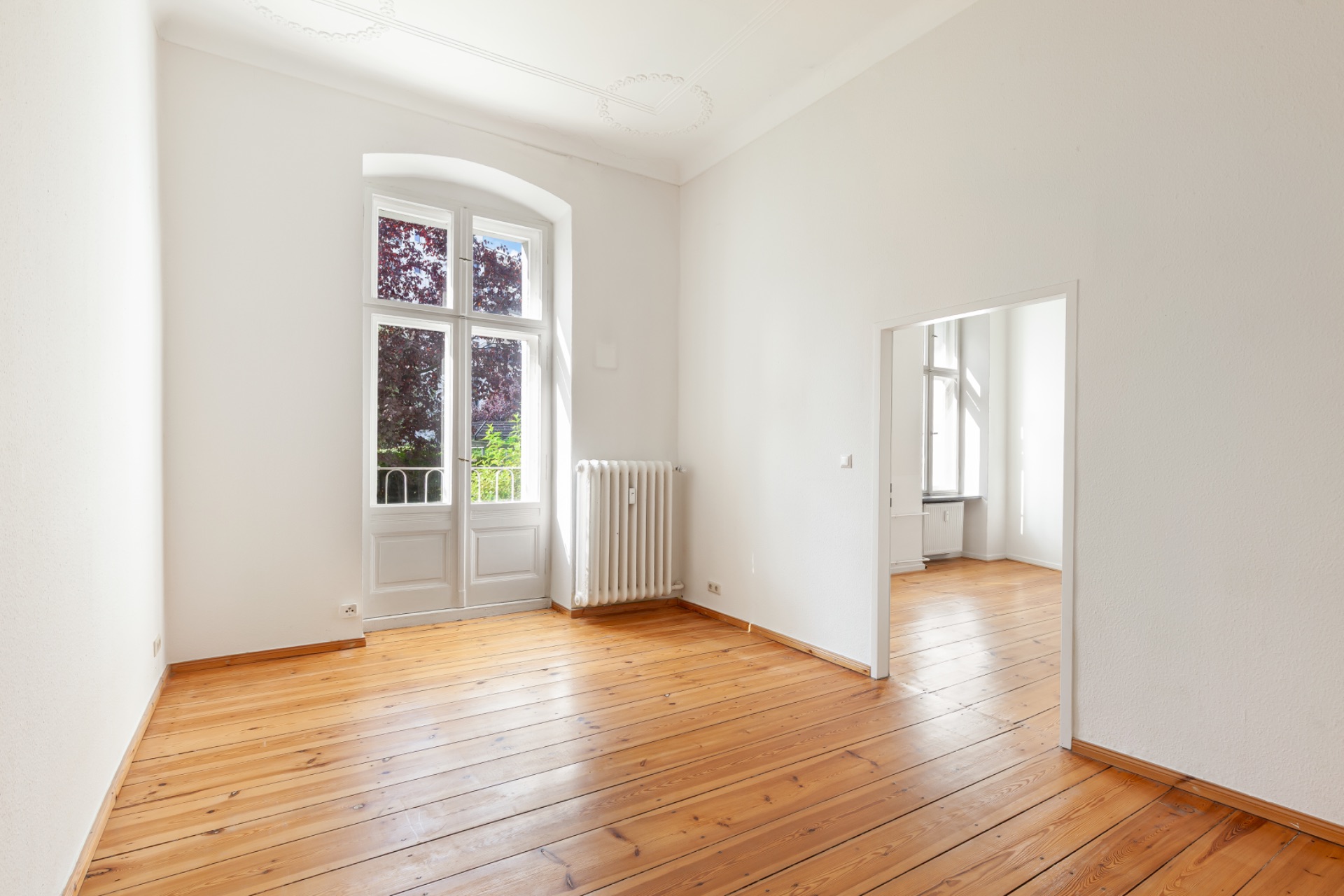 Helles Zimmer mit Holzboden, Fenstertür zum Balkon und Zugang zu weiterem Raum.