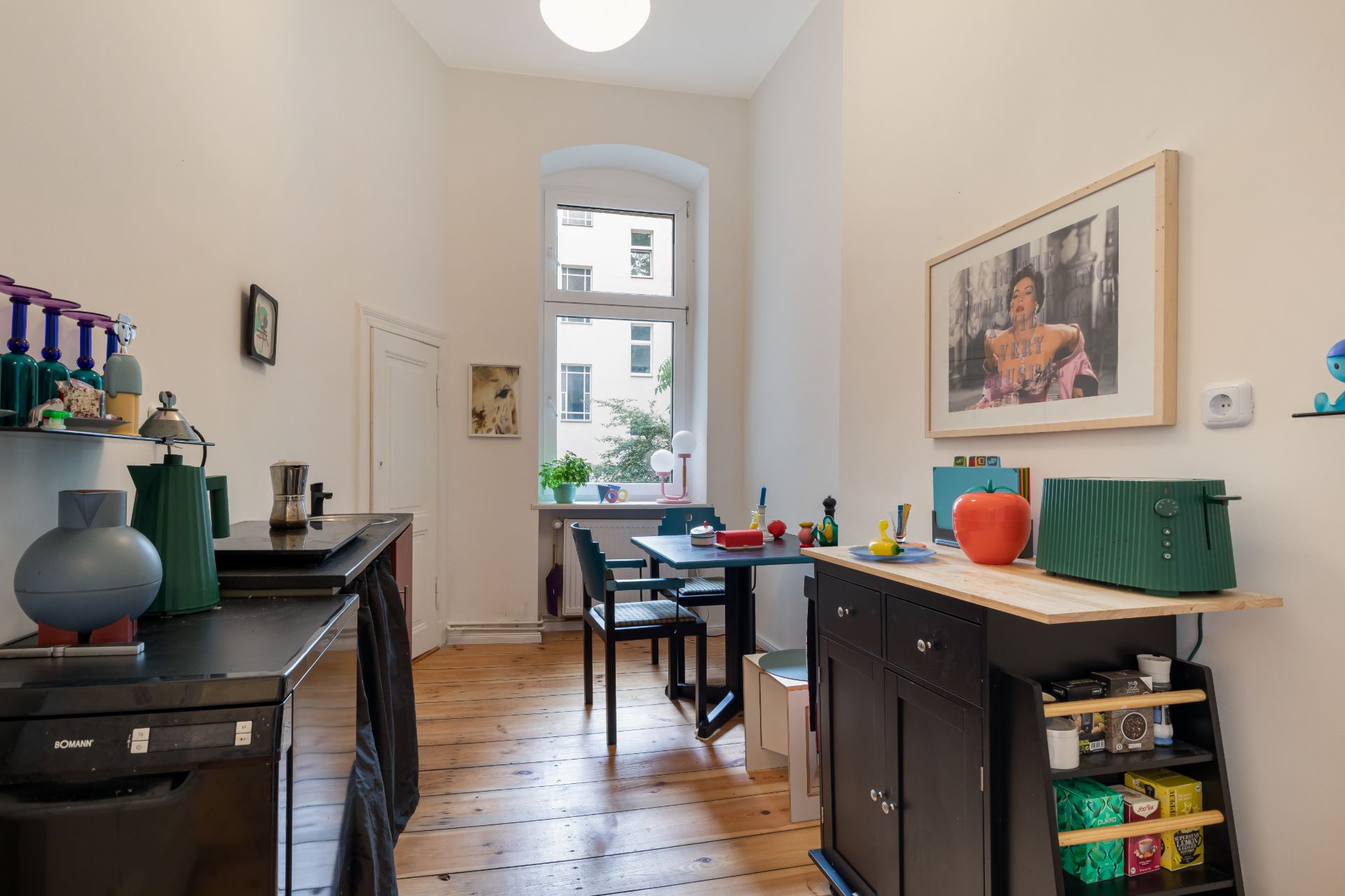 Helle Küche mit Holzboden, schwarzer Anrichte, Esstisch und Bild an der Wand.