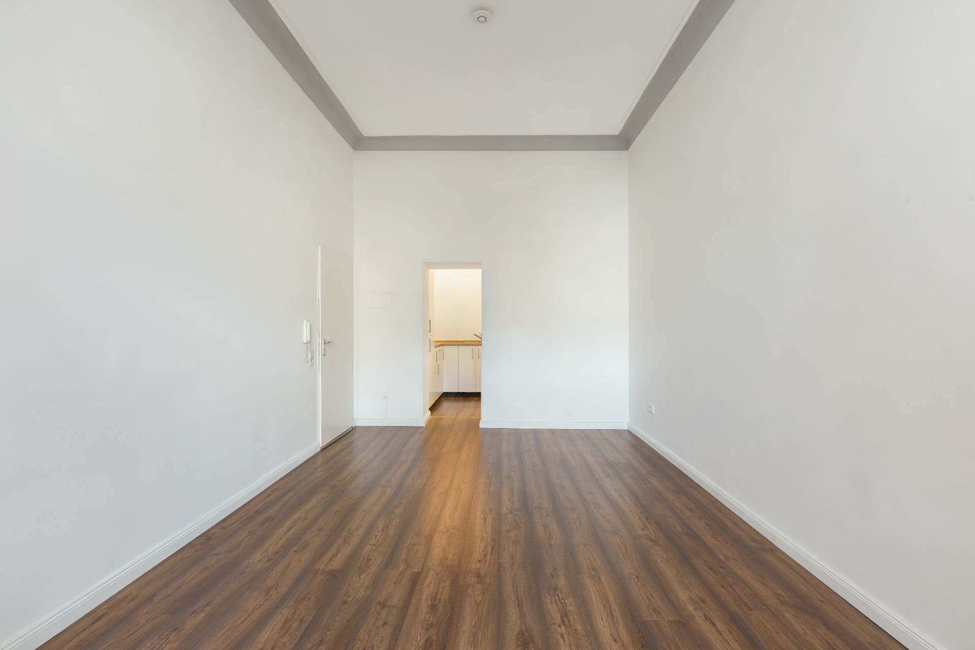 Leerer Raum mit weiß gestrichenen Wänden und braunem Holzboden, Tür führt in einen weiteren Raum.