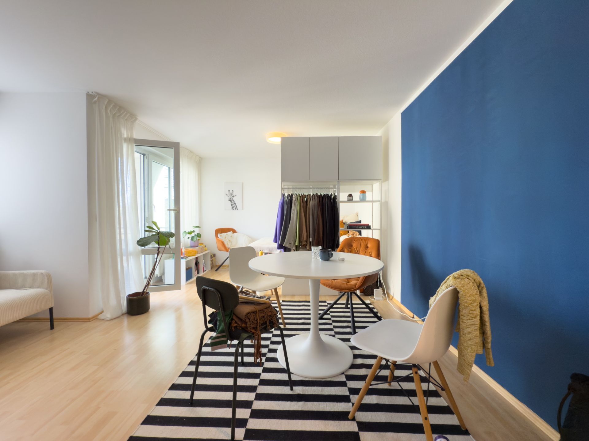 Helles Wohnzimmer mit rundem Tisch, Stühlen, blauem Wand, offener Garderobe und Pflanze.