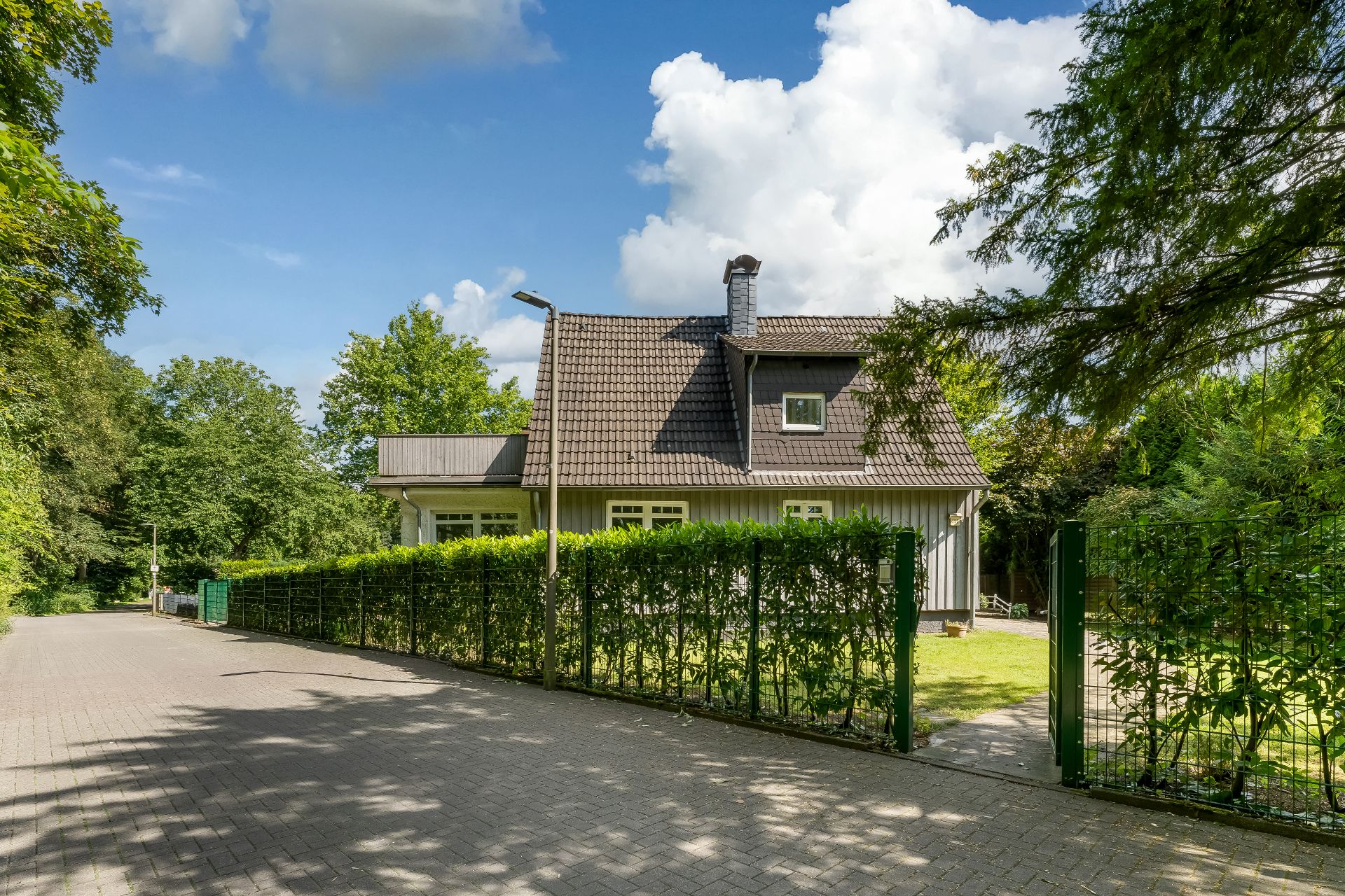 Einfamilienhaus mit Gartentor und Hecke, umgeben von Bäumen und blauem Himmel.