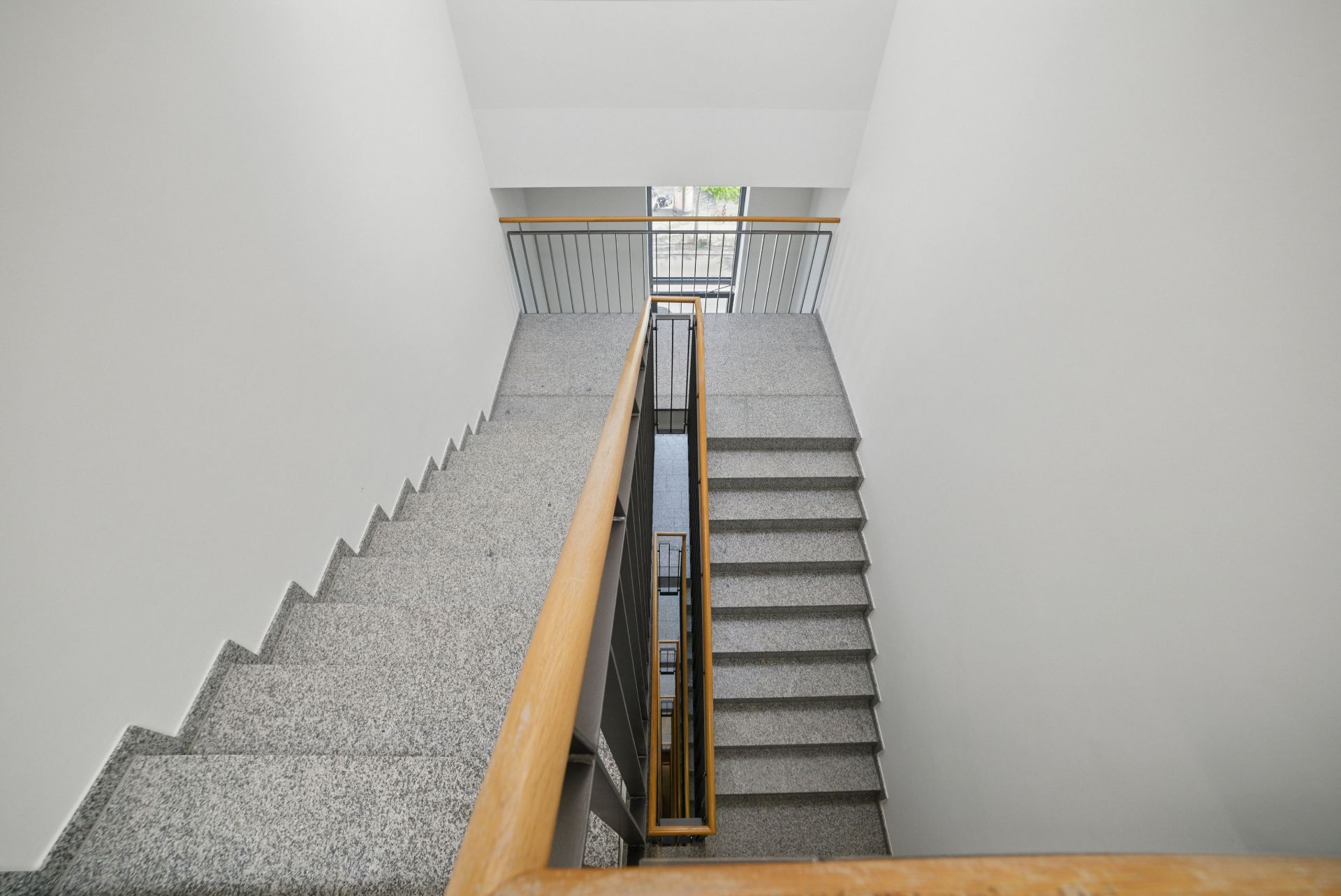 Treppe mit grauen Stufen und Holzgeländer in einem hellen Treppenhaus.