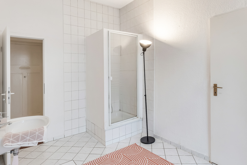 Helles Badezimmer mit Dusche, Waschbecken, Stehlampe und offenem Türrahmen.