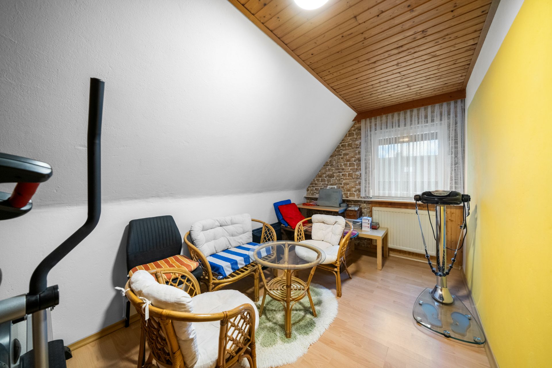 Kleines Zimmer mit Rattanstühlen, Glastisch, Sportgerät und Fenster mit Vorhang.