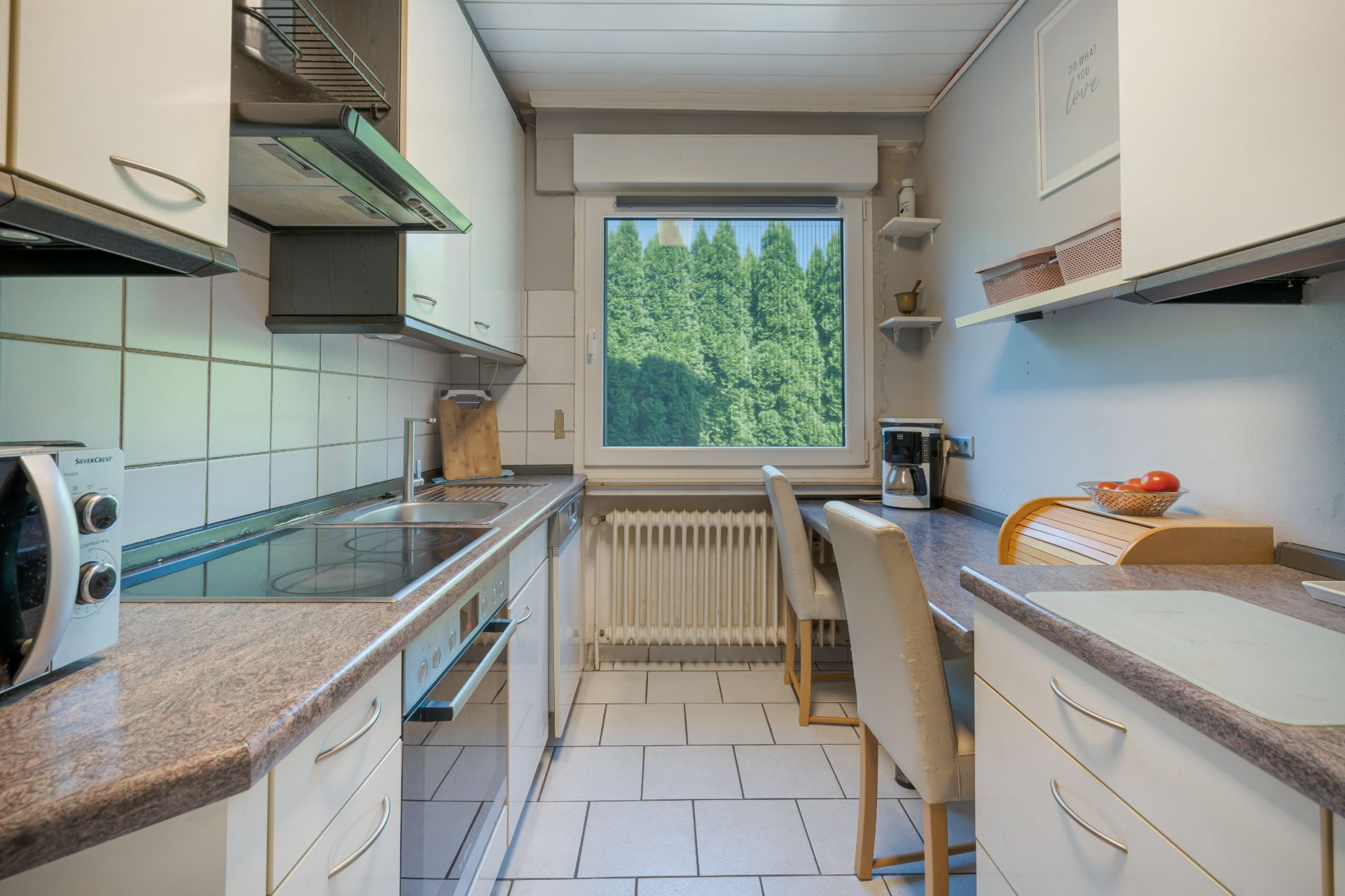 Helle Küche mit weißen Schränken, Esstisch und großem Fenster mit Blick auf grüne Bäume.