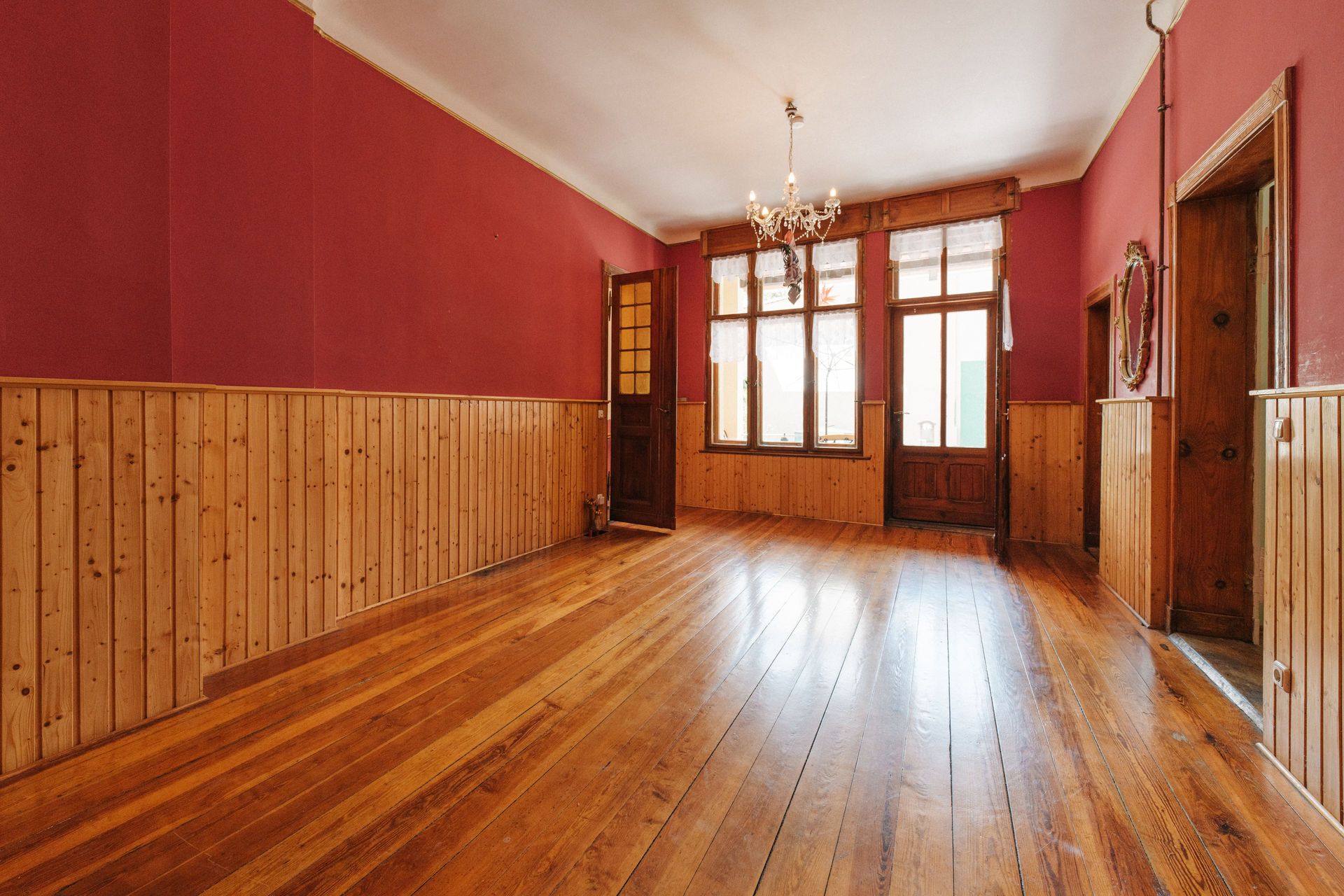 Leerer Raum mit Holzboden, roten Wänden, viel Fensterfläche und Kronleuchter an der Decke.