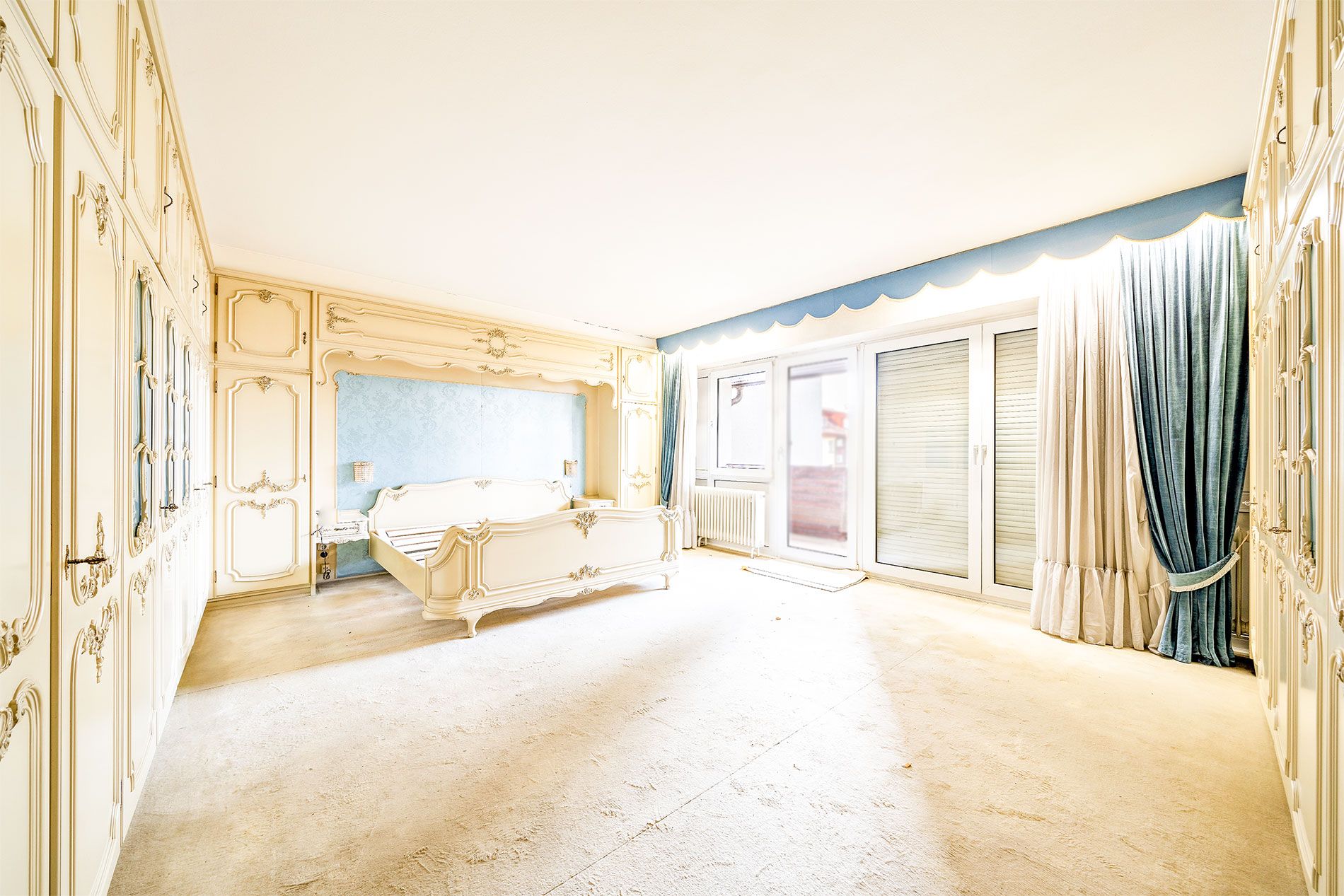 Helles Schlafzimmer mit cremefarbenen Schränken, Bett und großen Fenstern mit blauen Vorhängen.